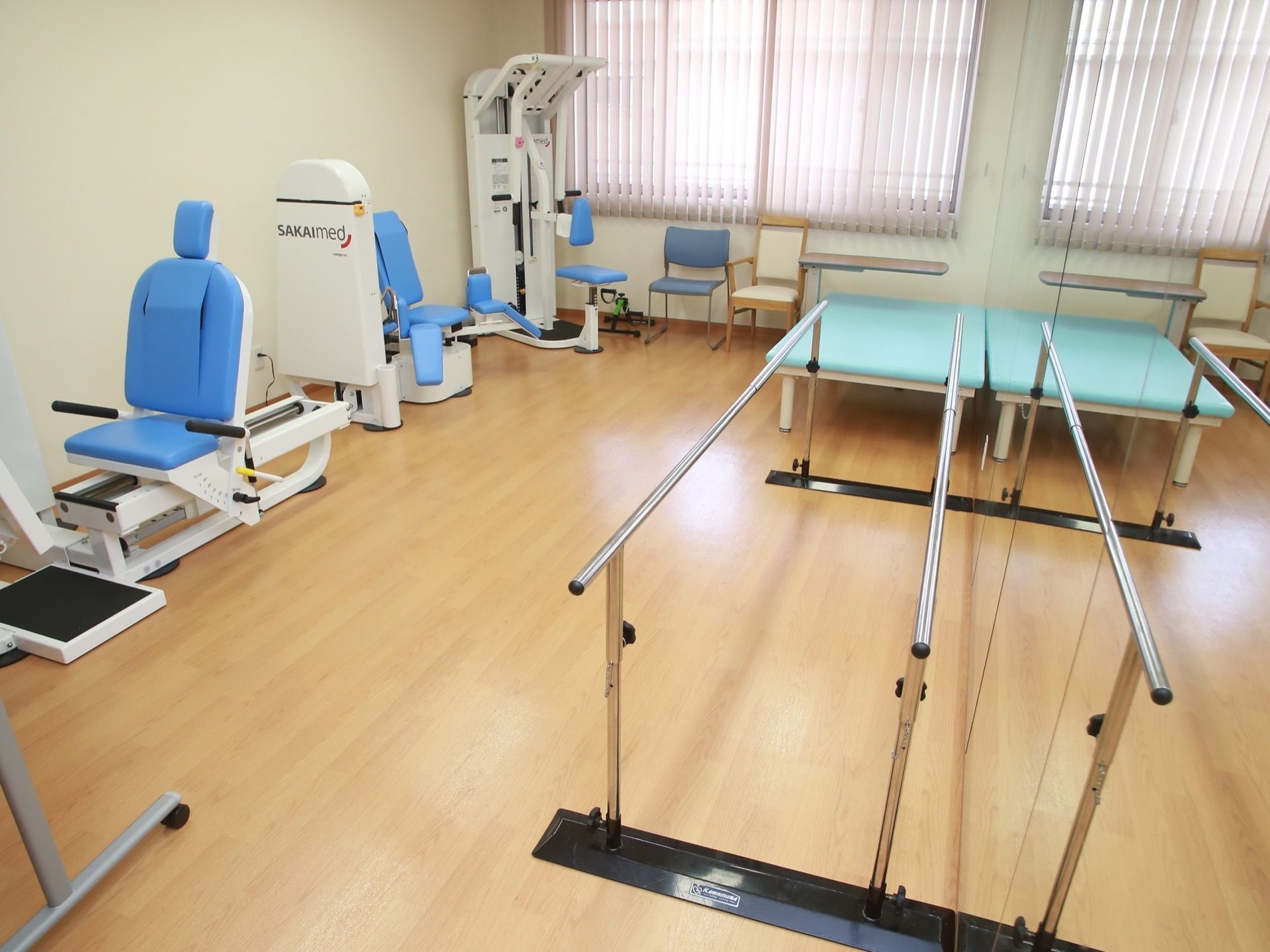 歩行訓練用の器具、筋力トレーニング用の器具などが整然と並べられている広間。窓際に椅子が複数用意されている。