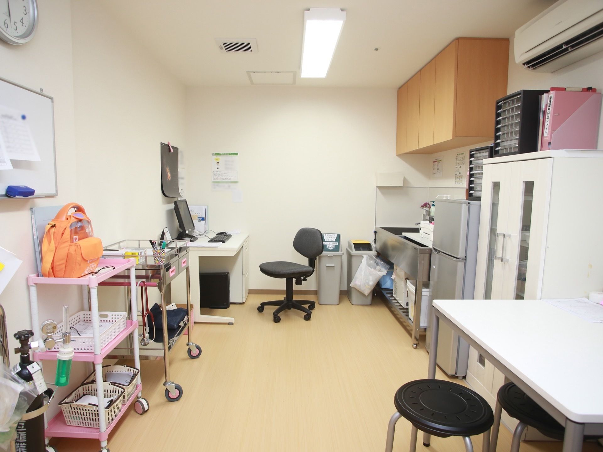 流し台の設置してある部屋に、冷蔵庫や棚が並んでいる。部屋の奧にデスクと椅子があり、手前にはテーブルと椅子が用意されている。