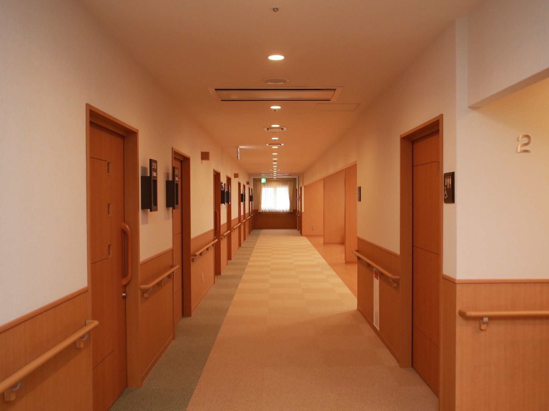 廊下の左側の壁に、居室の出入口が並んでいる。通路の突き当たりに窓があり、天井の照明も連続していて、明るい空間である。