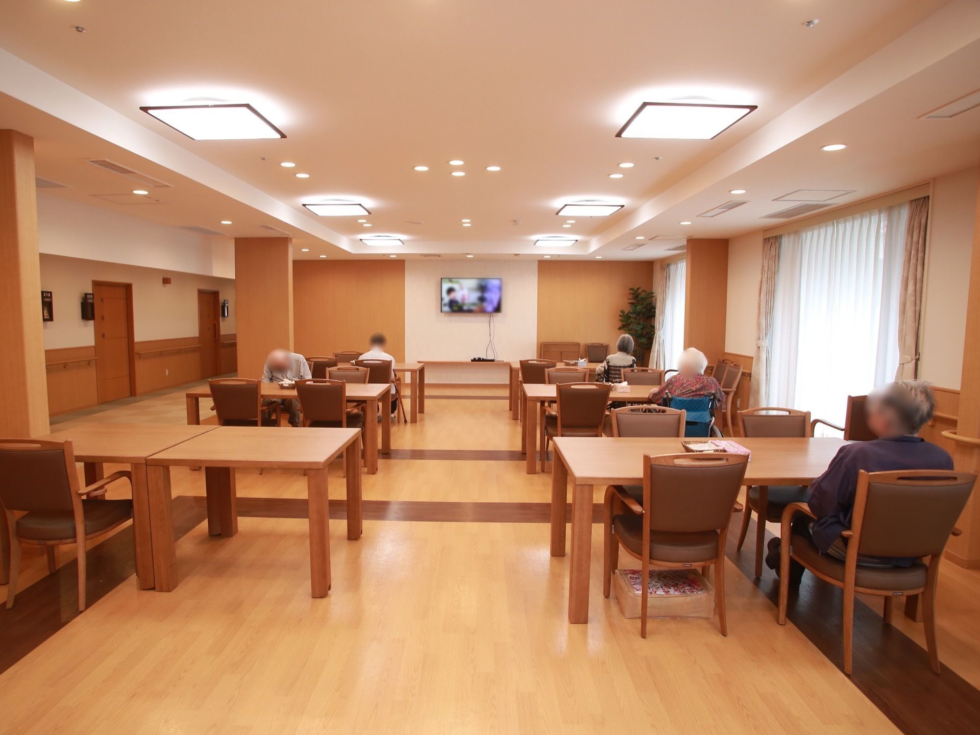 掃き出しの窓がある明るい空間に、ダイニングセットが整然と並べられている。大型テレビが壁の上方に設置されていて鑑賞しやすい。
