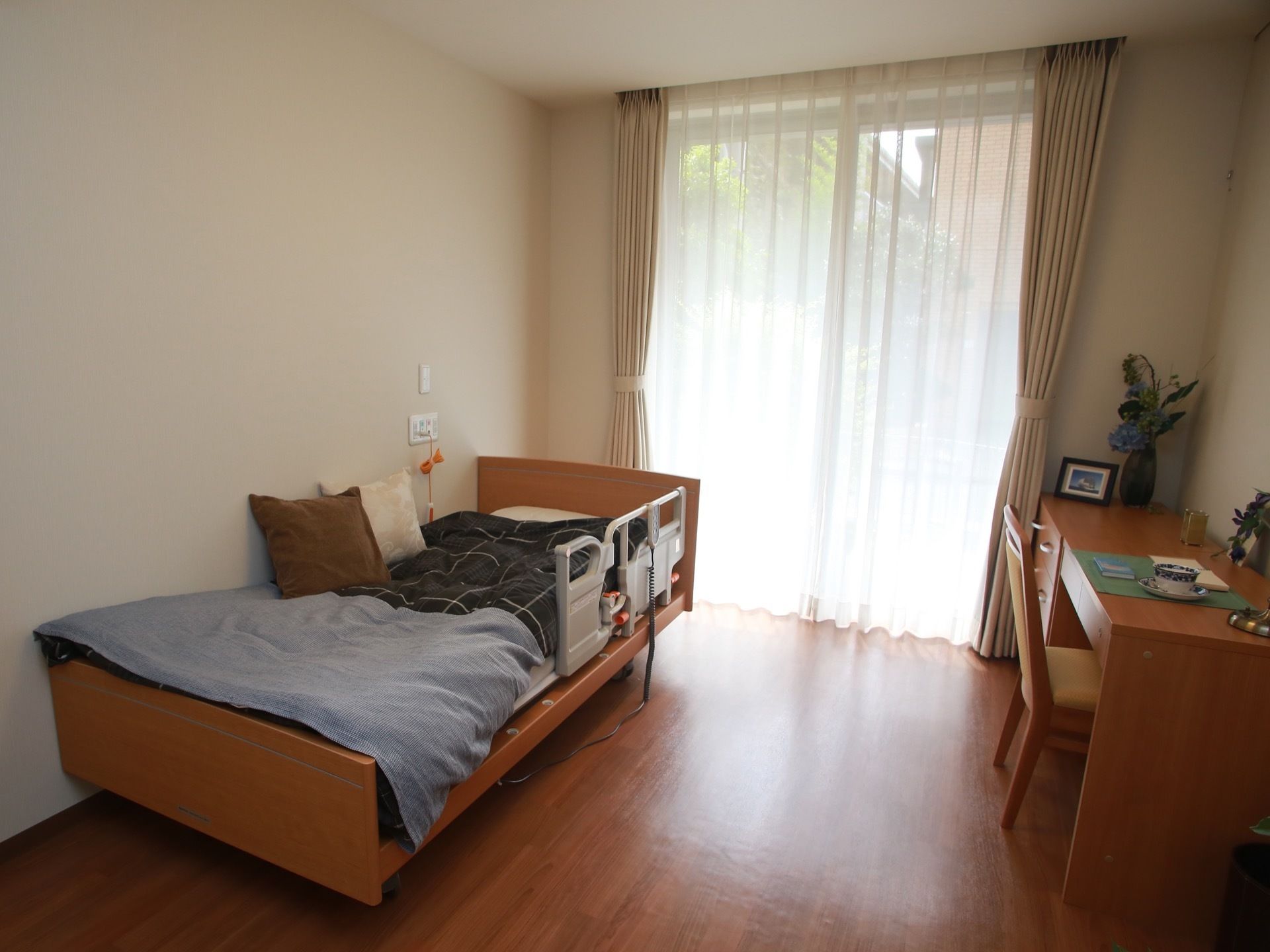 大きな窓のある部屋にベッドや机などの木製家具が置かれている。ベッドの脇に非常用ブザーが設置されている。