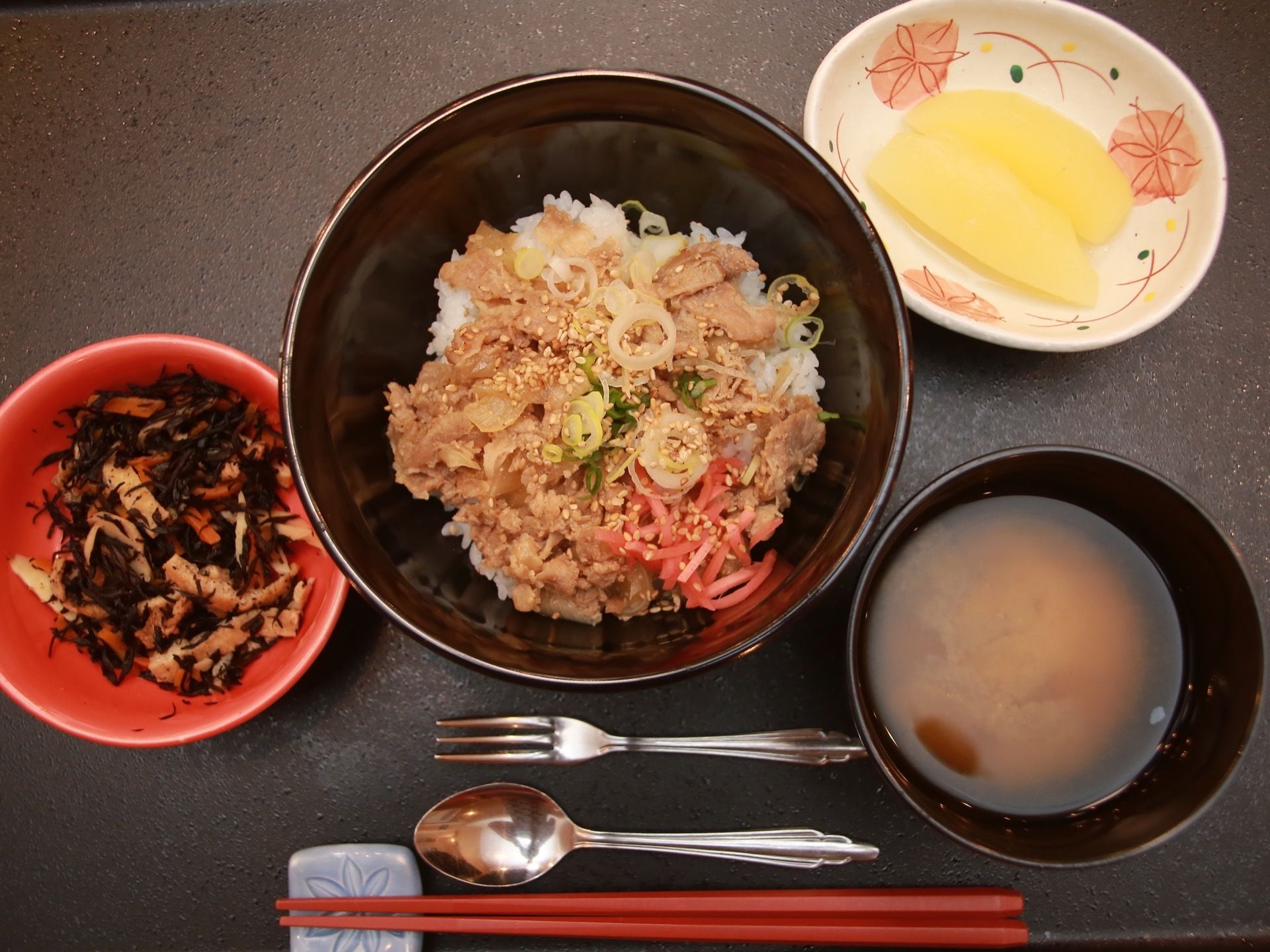 肉と野菜の丼物、味噌汁、五目煮、たくあん漬けがそれぞれ器に盛りつけられている。箸の他に、小さなフォークとスプーンが置かれている。