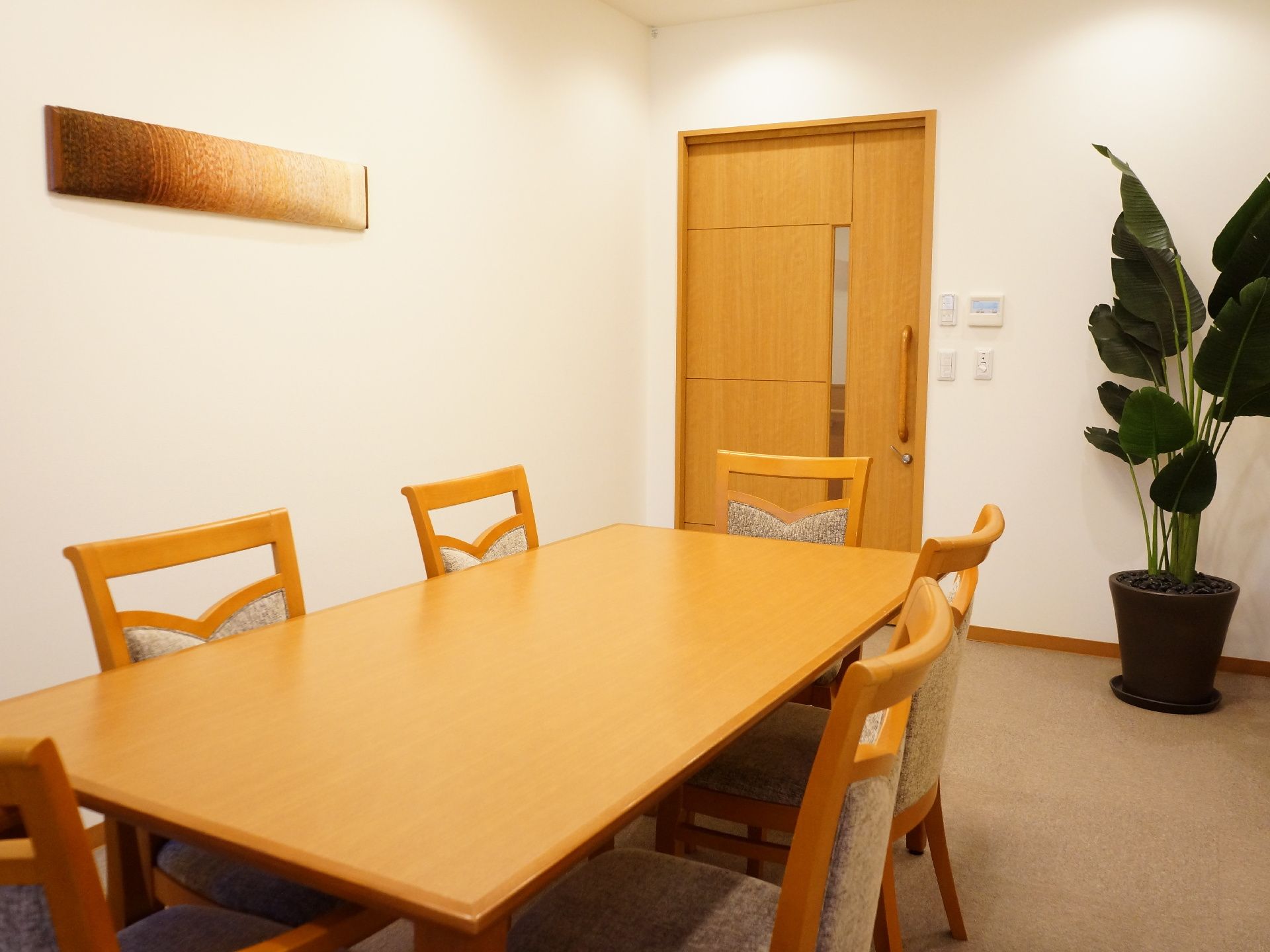 白い壁に木でできたドアがある。ドアには細長いガラスの部分がある。部屋の角に葉の大きい観葉植物が置いてある。テーブルと椅子がある。