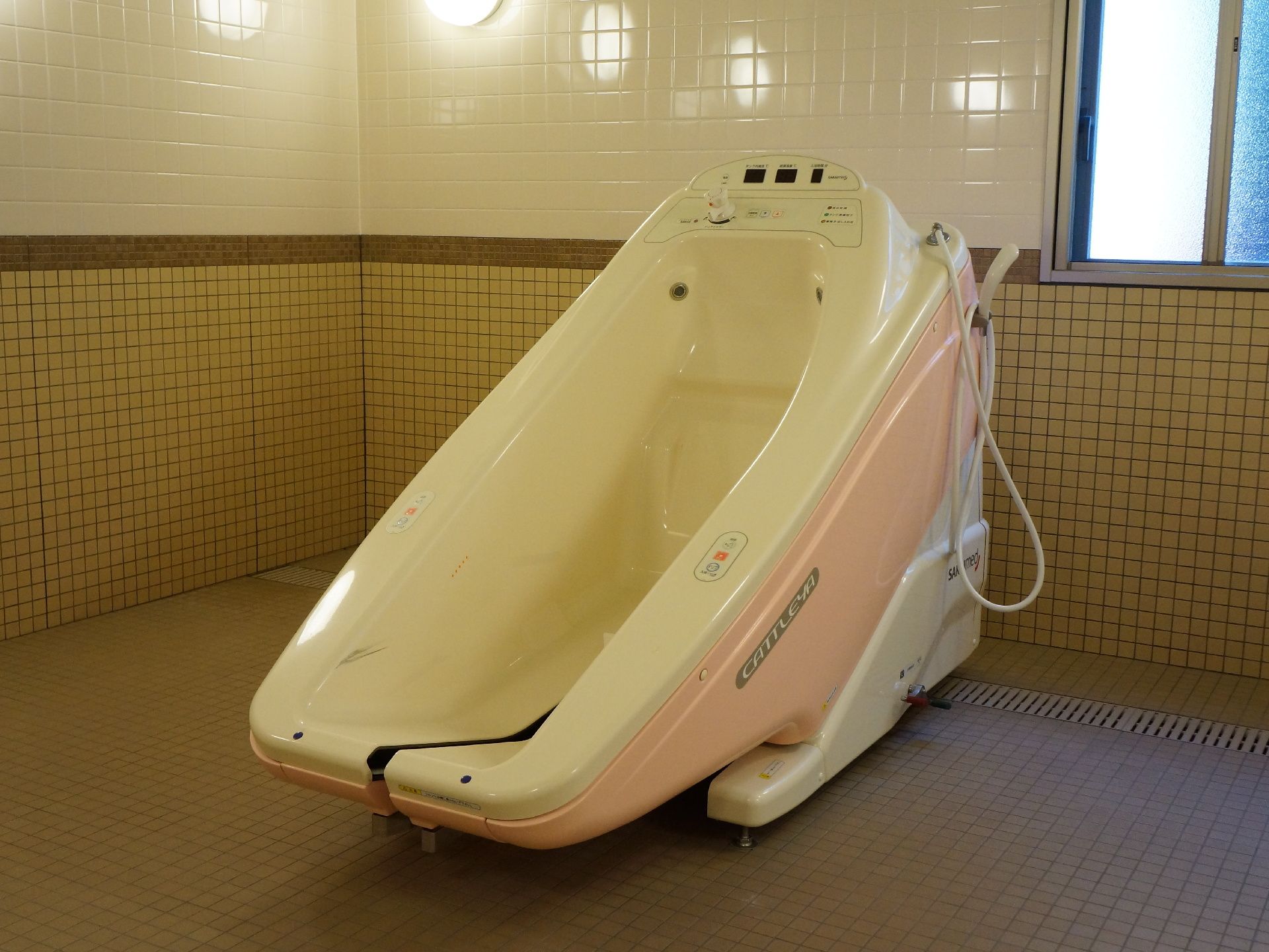 正面には入浴用介助器具が置かれている。奥の床には排水溝があり、そこの上側には窓がある。床や壁はタイル張りになっている。