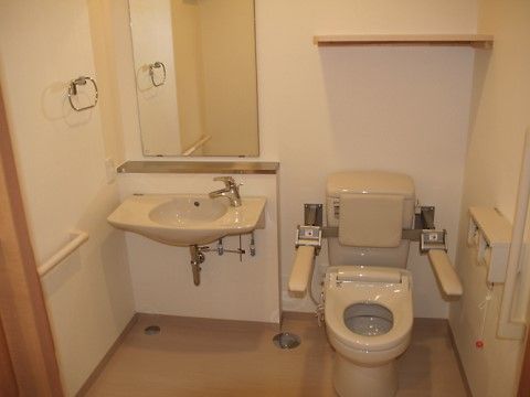 洗面台が完備されたトイレである。温水洗浄便座器には、可動式の手すりや背もたれパッドが設置され、安全にご利用いただける。