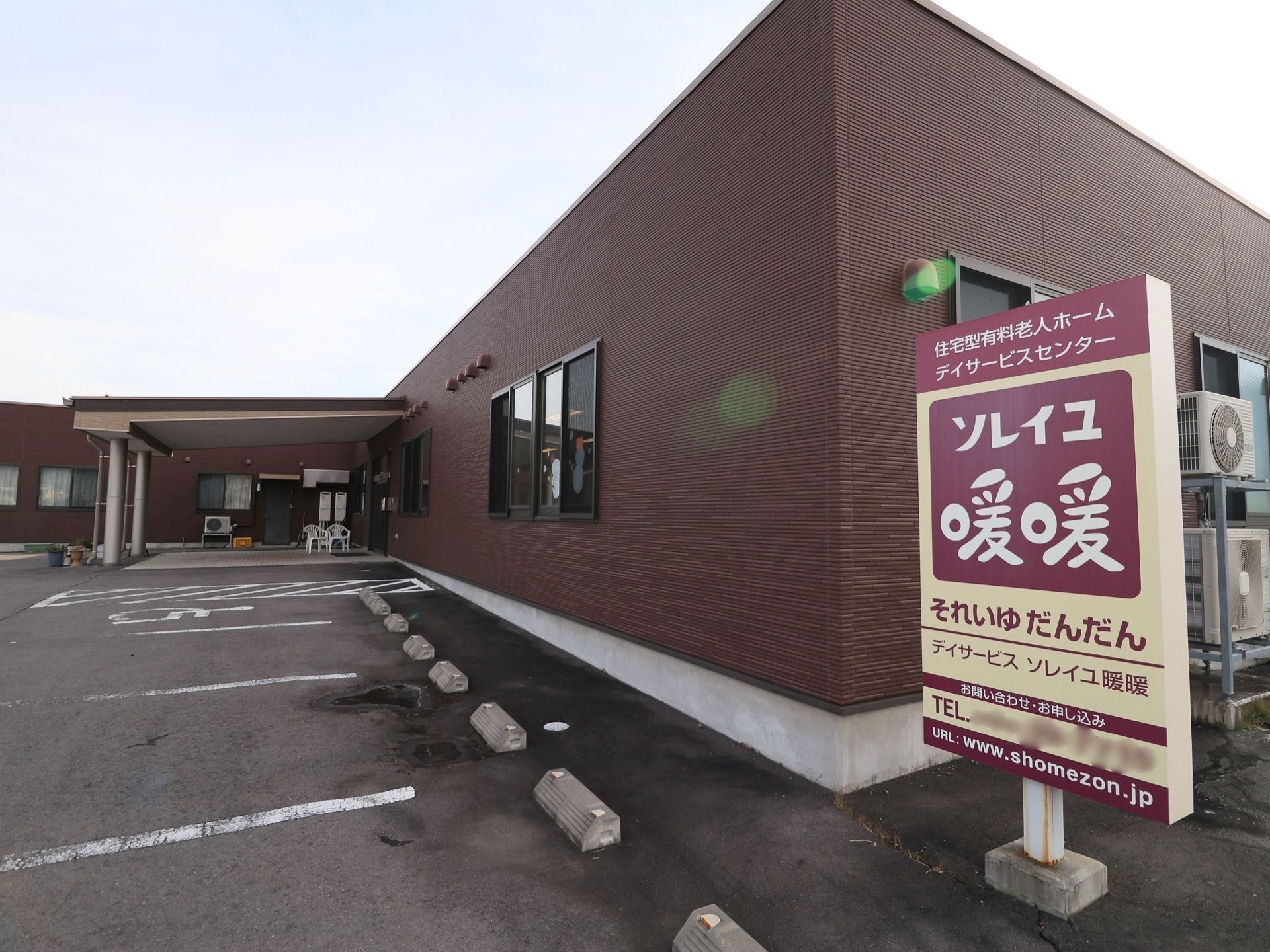 建物に隣接して、駐車場スペースが造られている。建物の隅には、室外機とともに施設名が書かれた看板が設置されている。