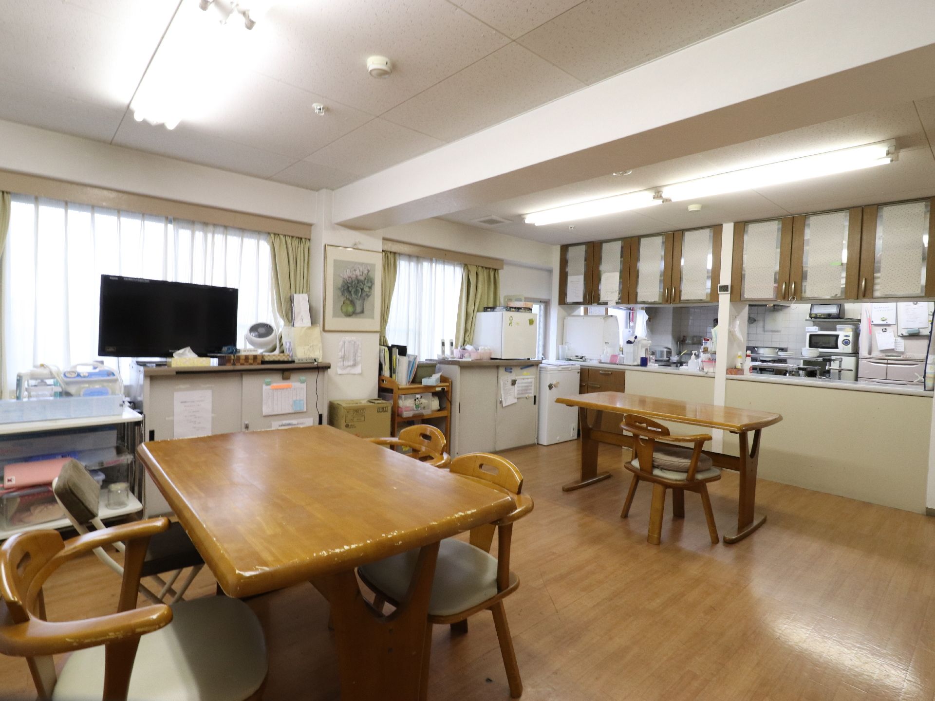 台所が見える食堂兼多目的室で、窓そばに一台テレビが置いてある。木製のテーブルが二台ある。冷蔵庫が大小置いてある。