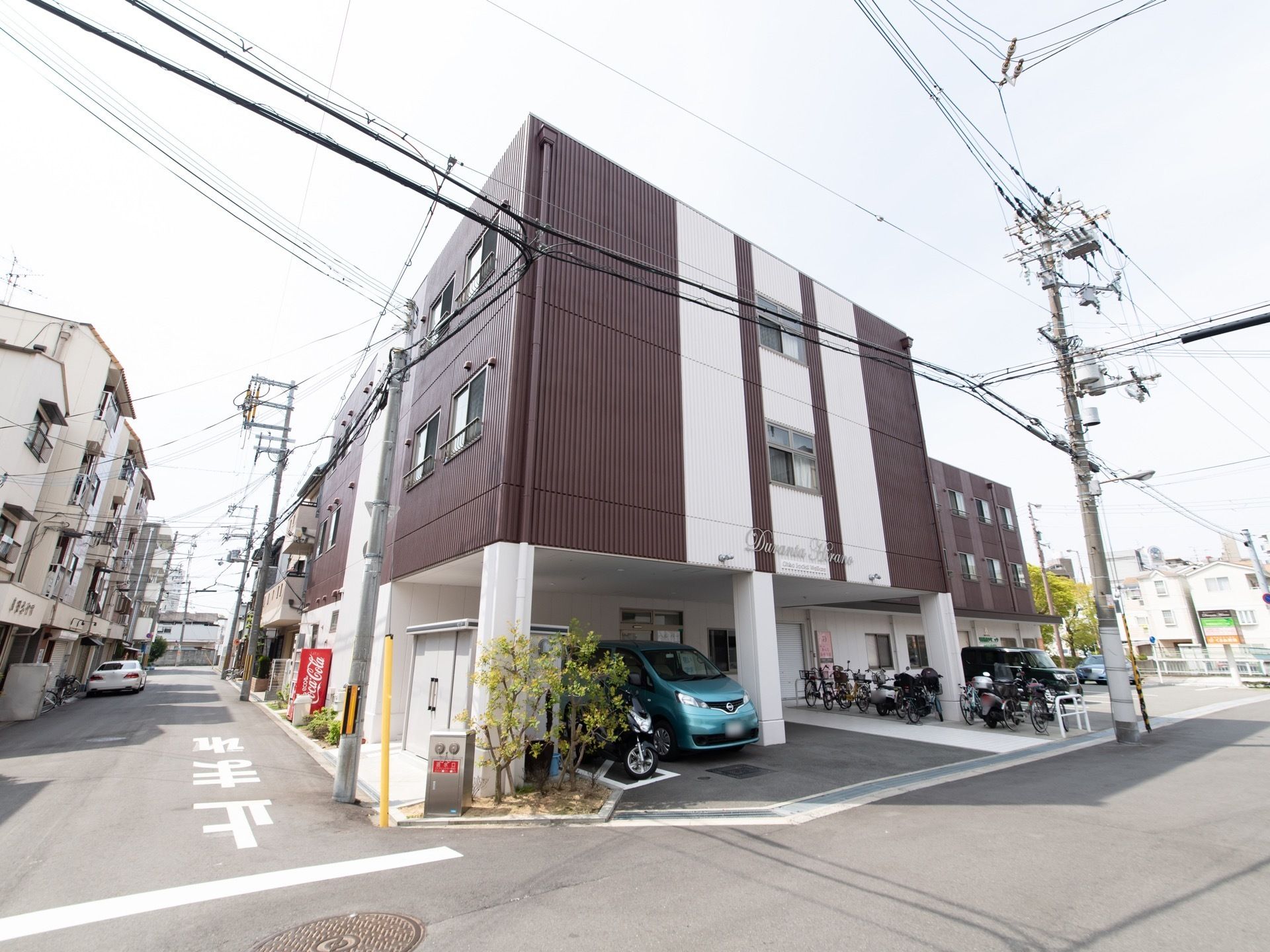 白と茶色を基調としたモダンな造りの建物である。住宅街の角地に建てられていて、１階部分に車が停まっている。