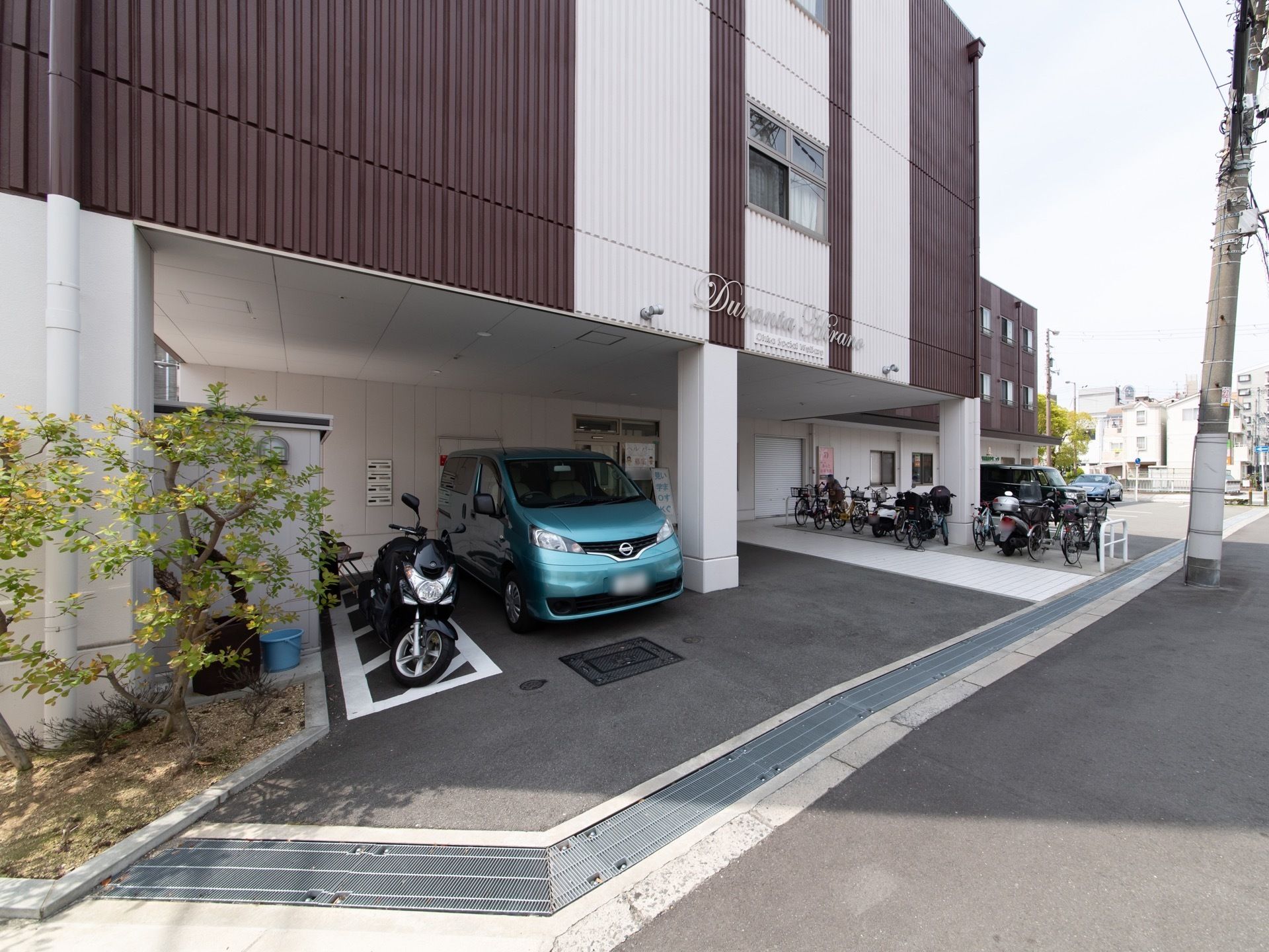 隅に木が植えられている。建物の一部の１階に車が停められている。駐輪場が設置されていて、自転車が置かれている。