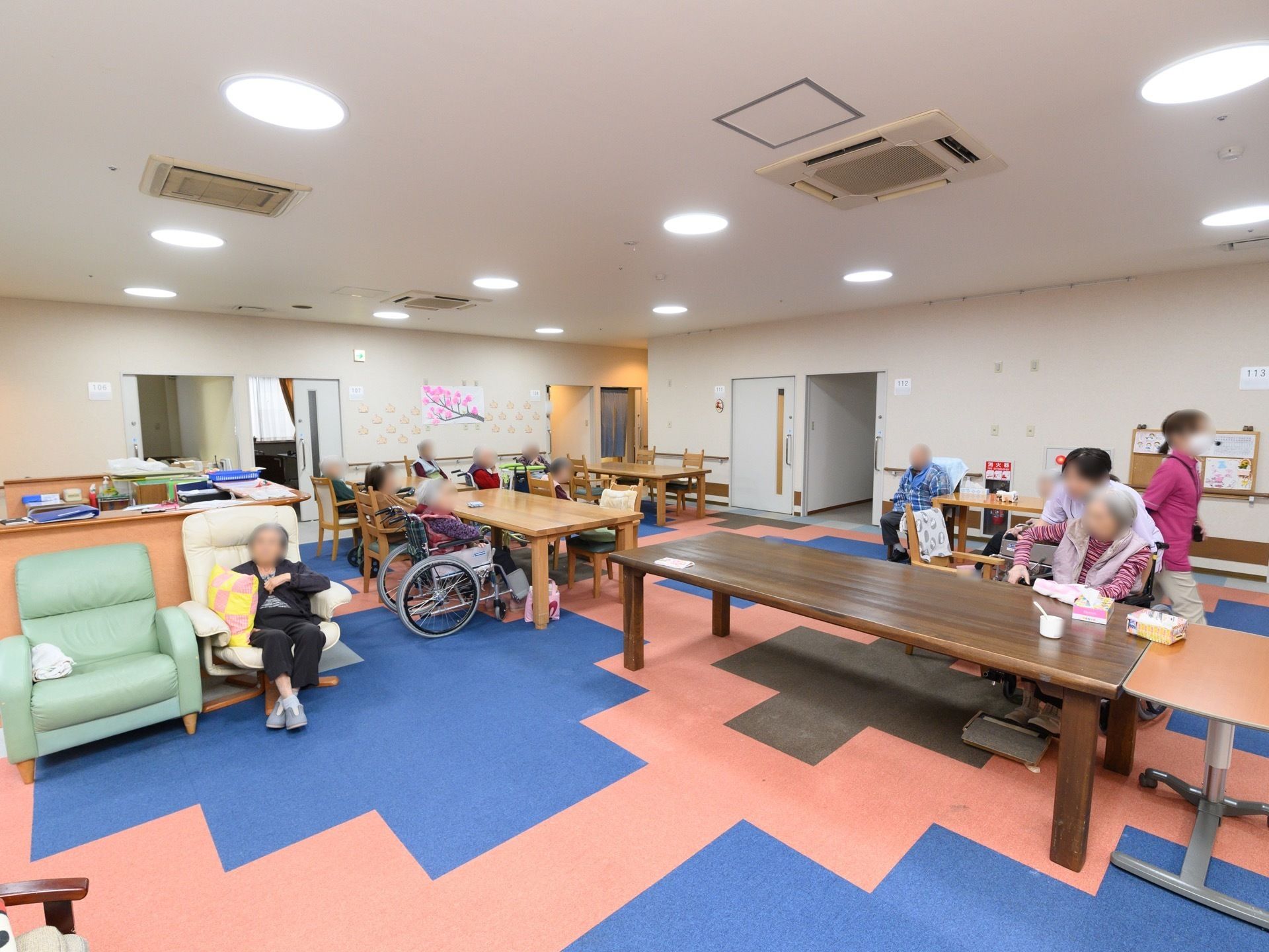 カラフルな床面の共有スペースがある。女性スタッフが、車椅子をご利用の入居者様がテーブルにつく際の介助をしている。
