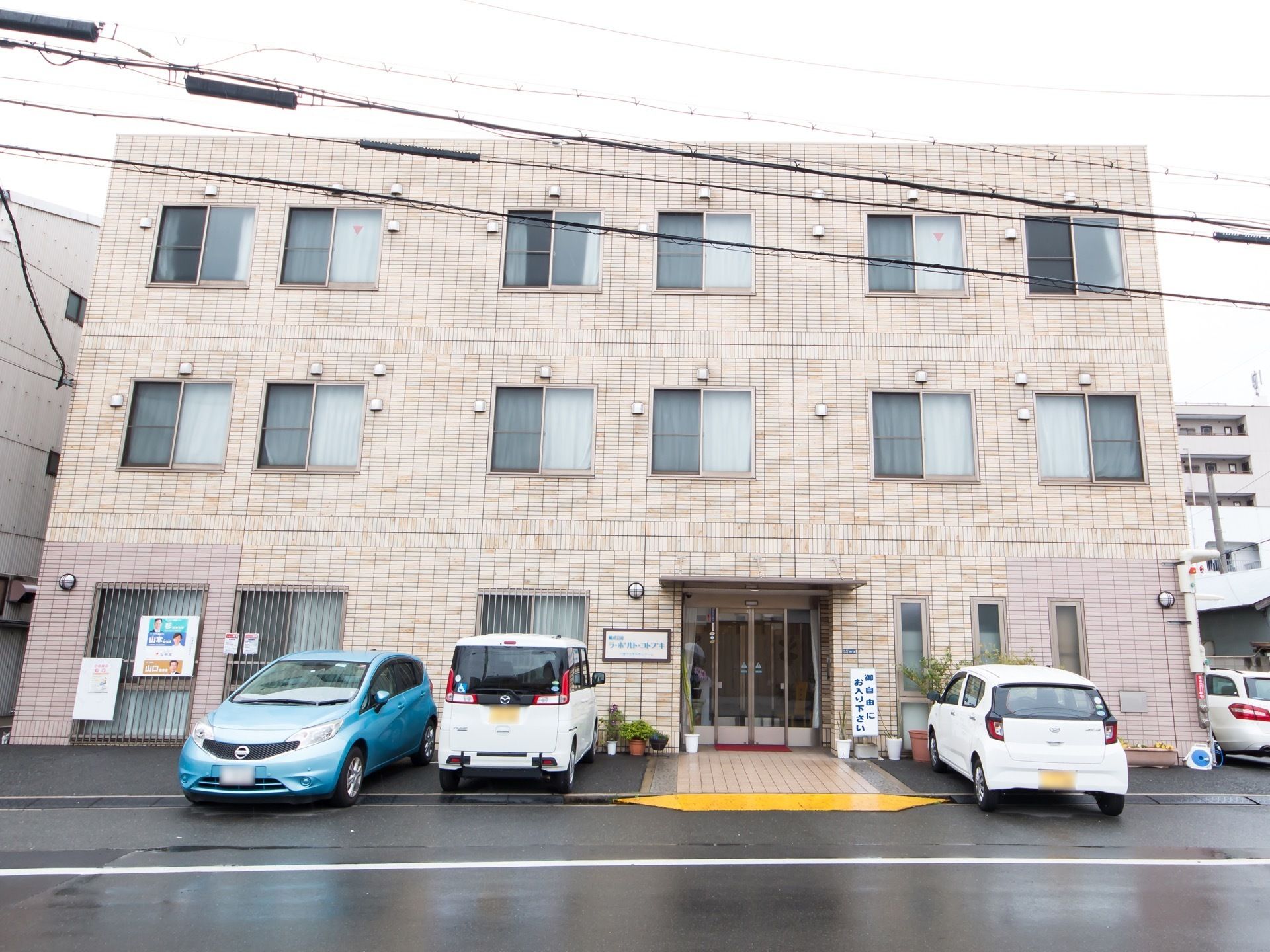 広い道路に面している３階建ての建物で、施設目の前の歩道にはスロープが設置されている。玄関横の外壁には、表札が取りつけられている。