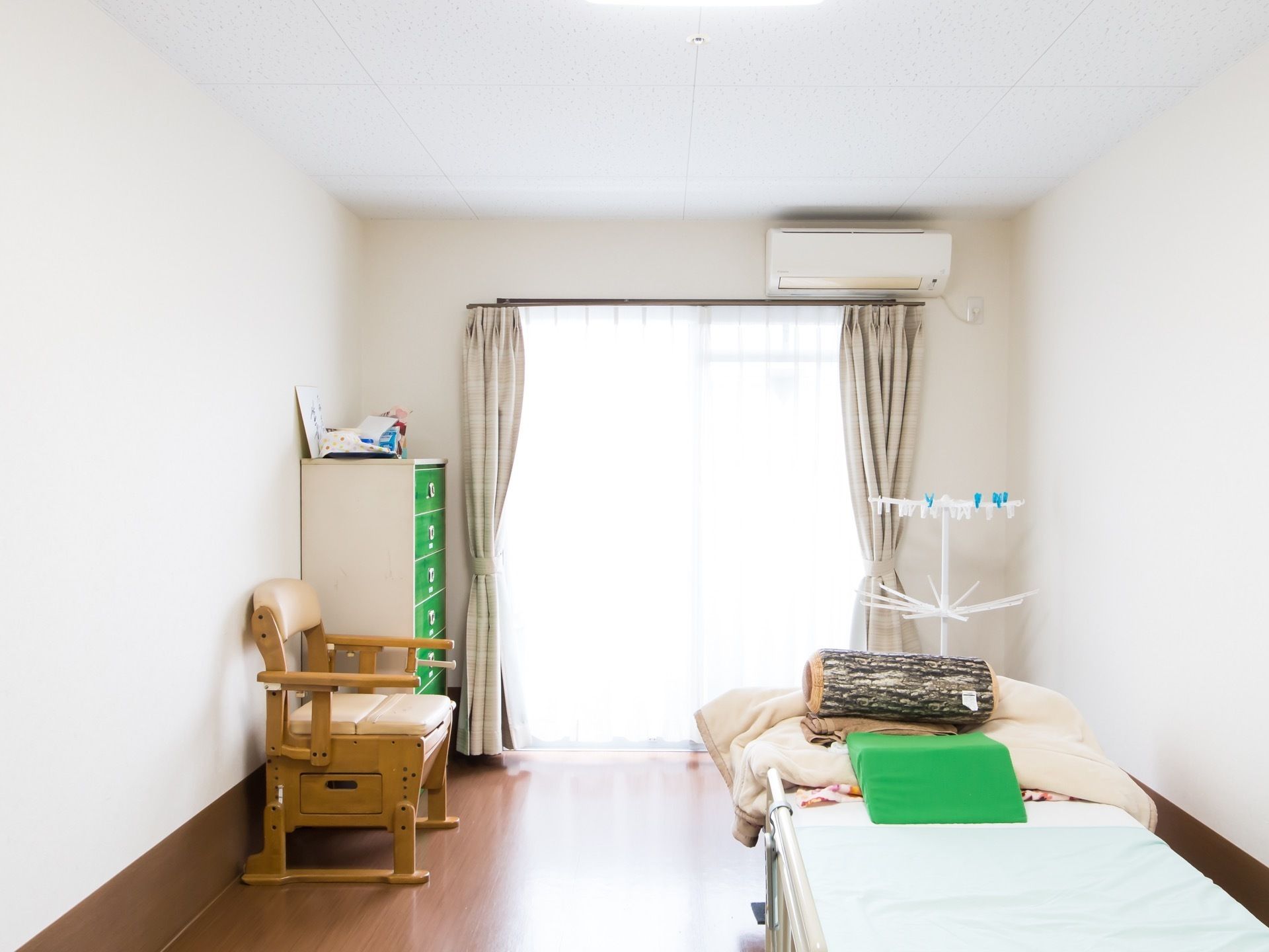 居室内の左手の壁に沿って緑の引き出しがはまったチェストと、木製の椅子が置かれている。右手にはベッドと物干し台がある。