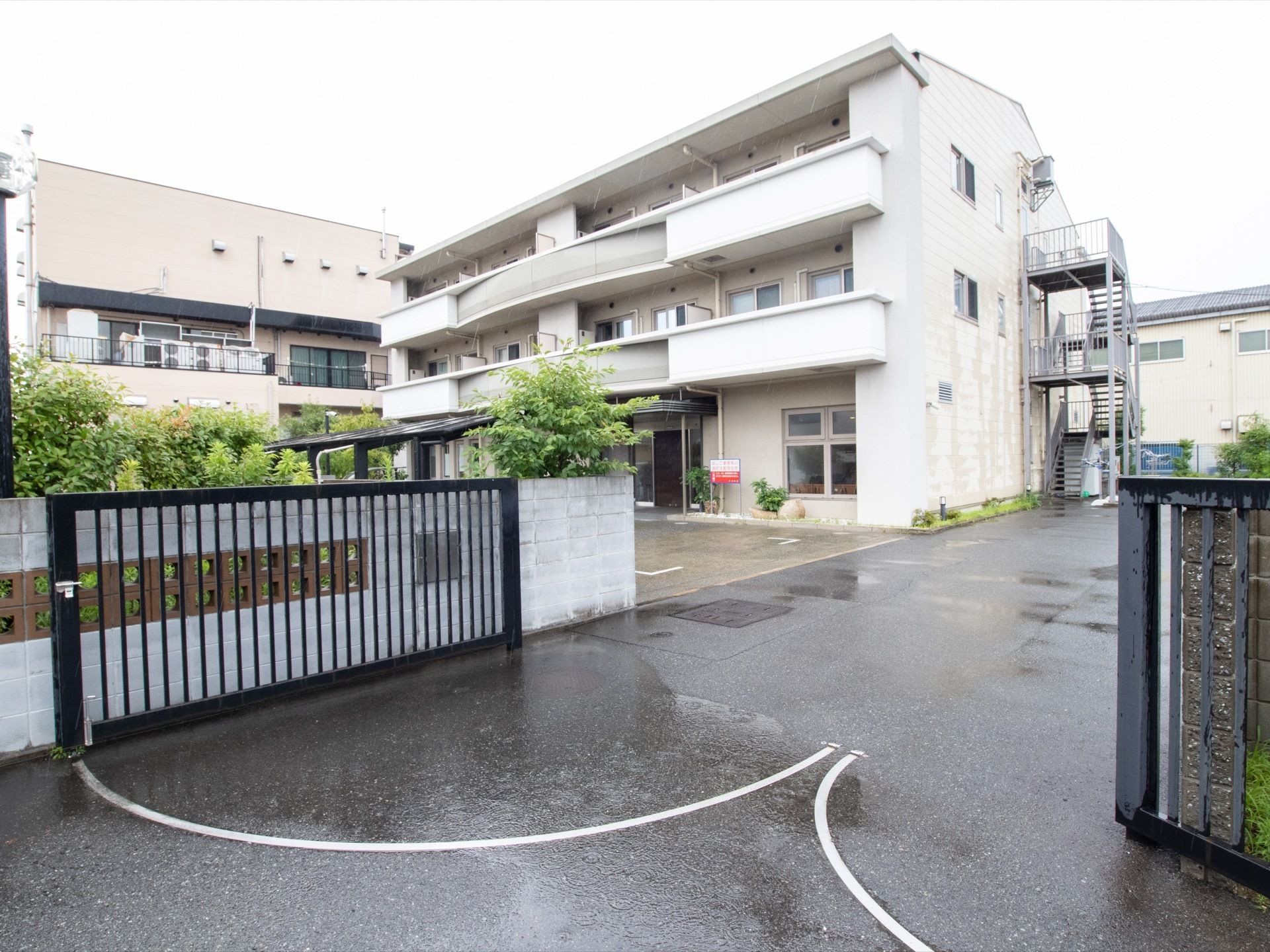 金属製の黒い大きな門扉とブロック塀で囲まれた広い敷地に、緑豊かな庭や駐車場が広がっている。その奧に３階建ての施設がある。