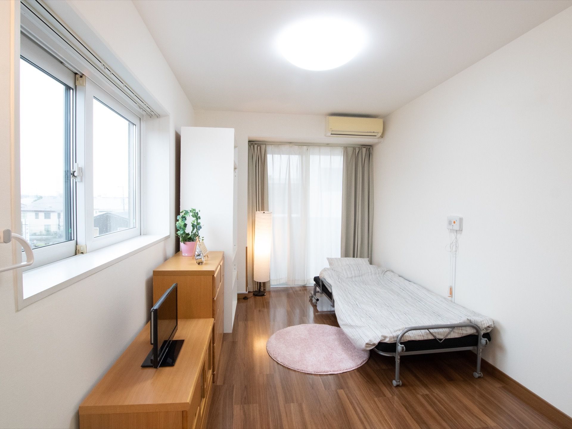 白い壁と濃い茶色のフローリングの部屋。窓が２面にあり、天井に大きな照明が設置され、枕元には間接照明もあり、とても明るい。