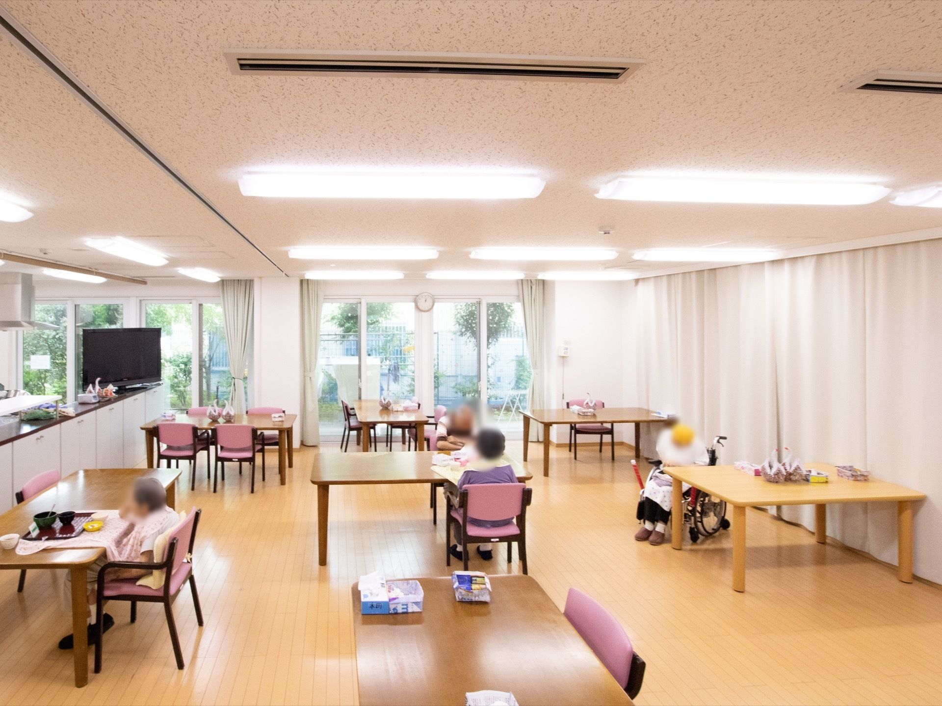 薄茶色のフローリングの広間に、大きな窓が並んでおり、広々と明るい印象の食堂である。木製テーブルが十分な間隔を空けて配置されている。