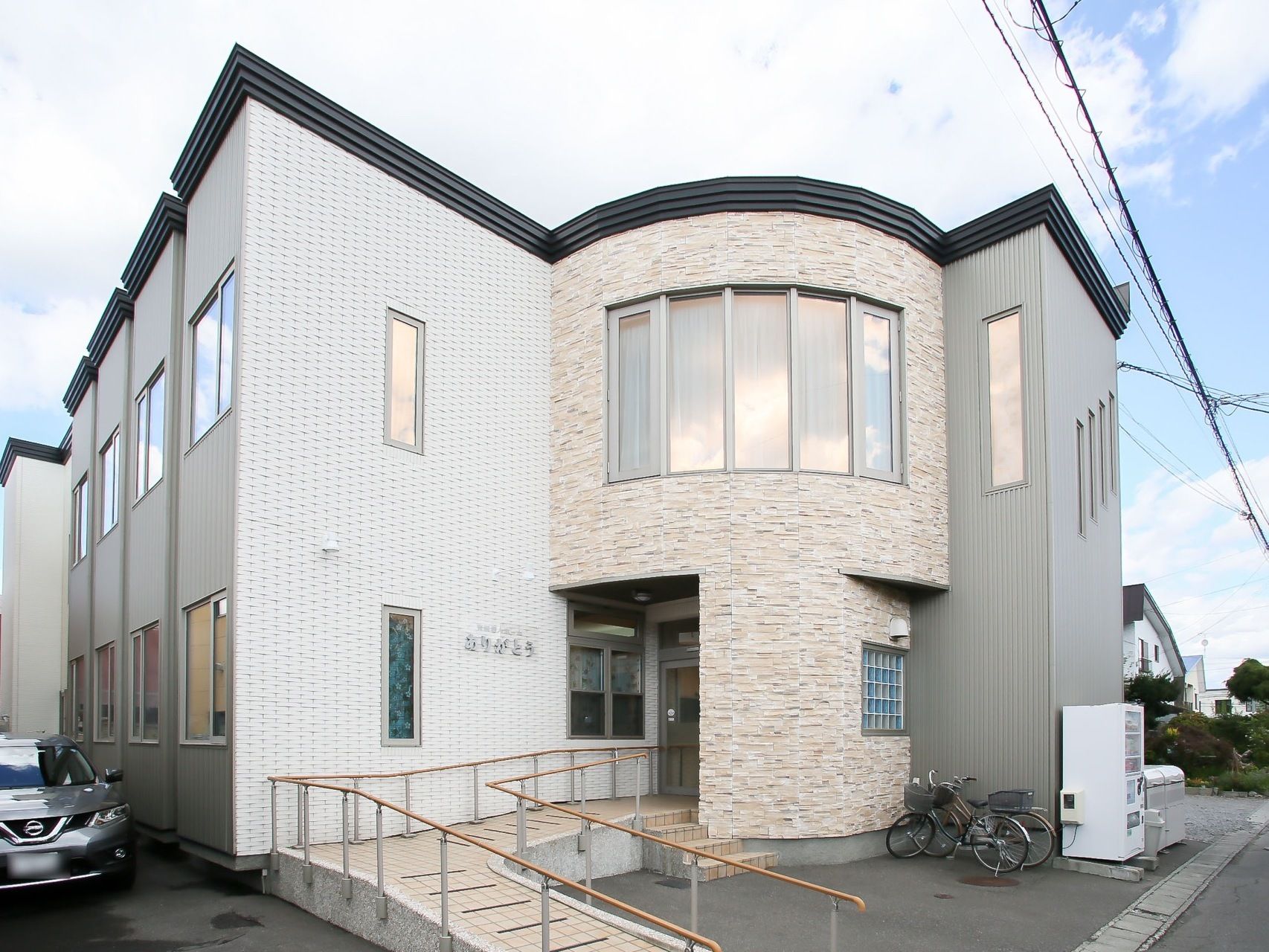 四角い建物の一部に円形のフロアがある。施設の外に自動販売機があり、その横にごみ箱が置かれ、自転車が２台並んでいる。