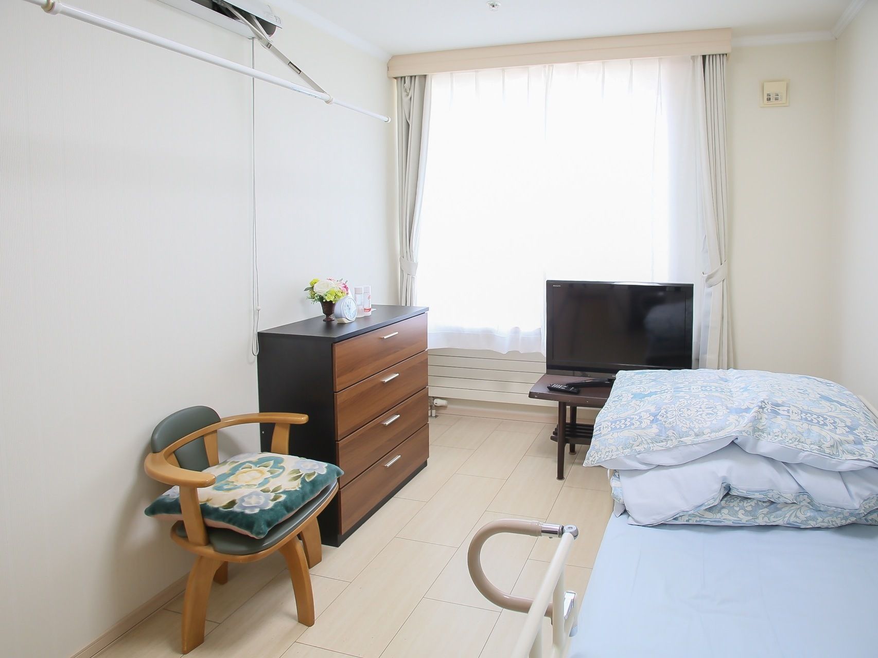 居室の壁際に介護用ベッドが置かれている。４段のチェストが備えつけられ、その横に椅子が１脚ある。窓の前に液晶テレビがある。