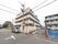 玄関正面側と横側は道路に隣接している。建物傍には電柱があり、車が1台停まっている。周辺は住宅やアパートが建ち並んでいる。