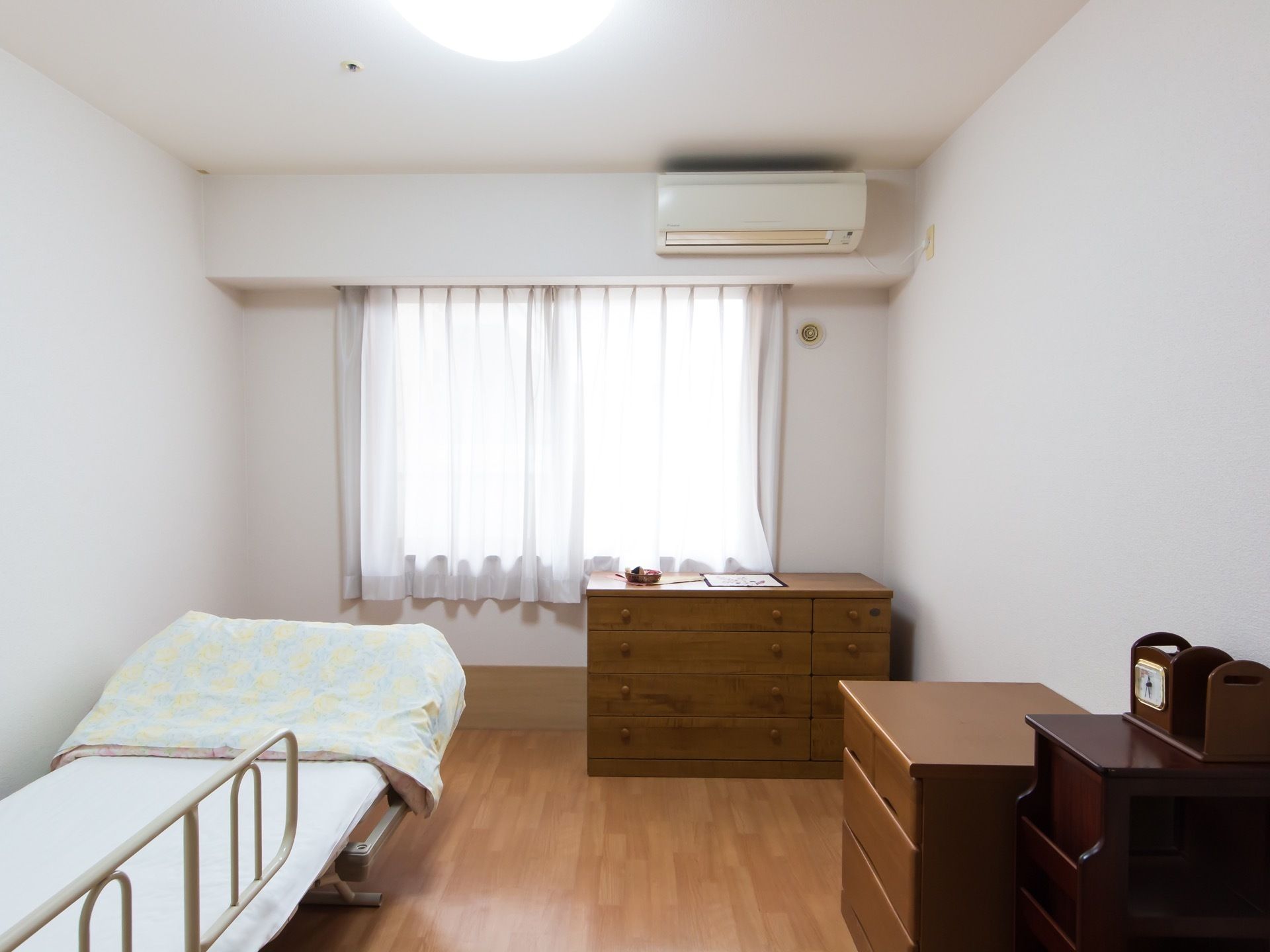 木目調の床に、壁と天井は白色のクロスである。カーテンが掛かった腰高窓の上にエアコンが設置されている。壁際にベッドがある。