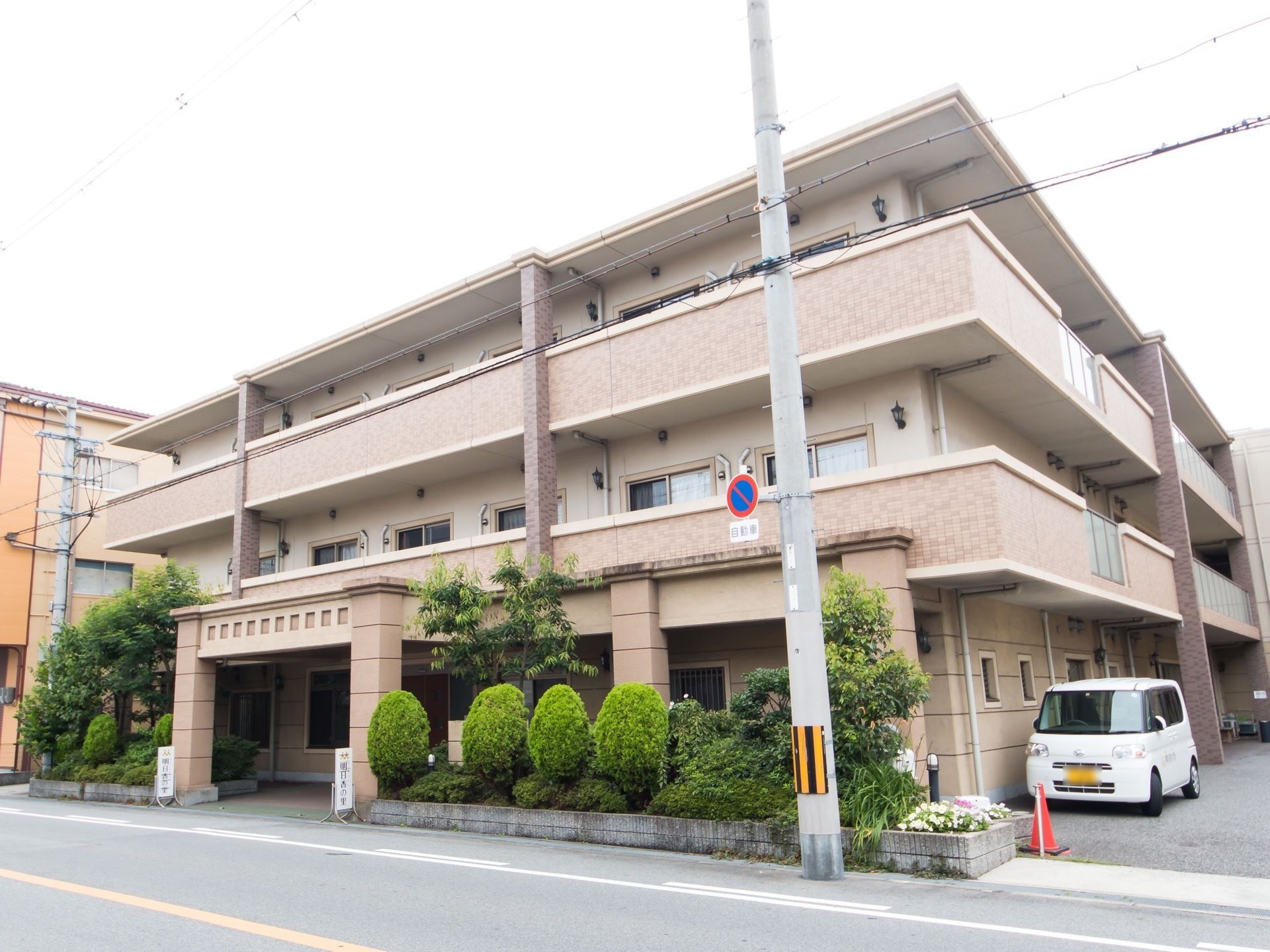 施設前の道路に面して植栽帯がある。建物の横が車の乗り入れ口になっており、車が建物に沿って停められている。