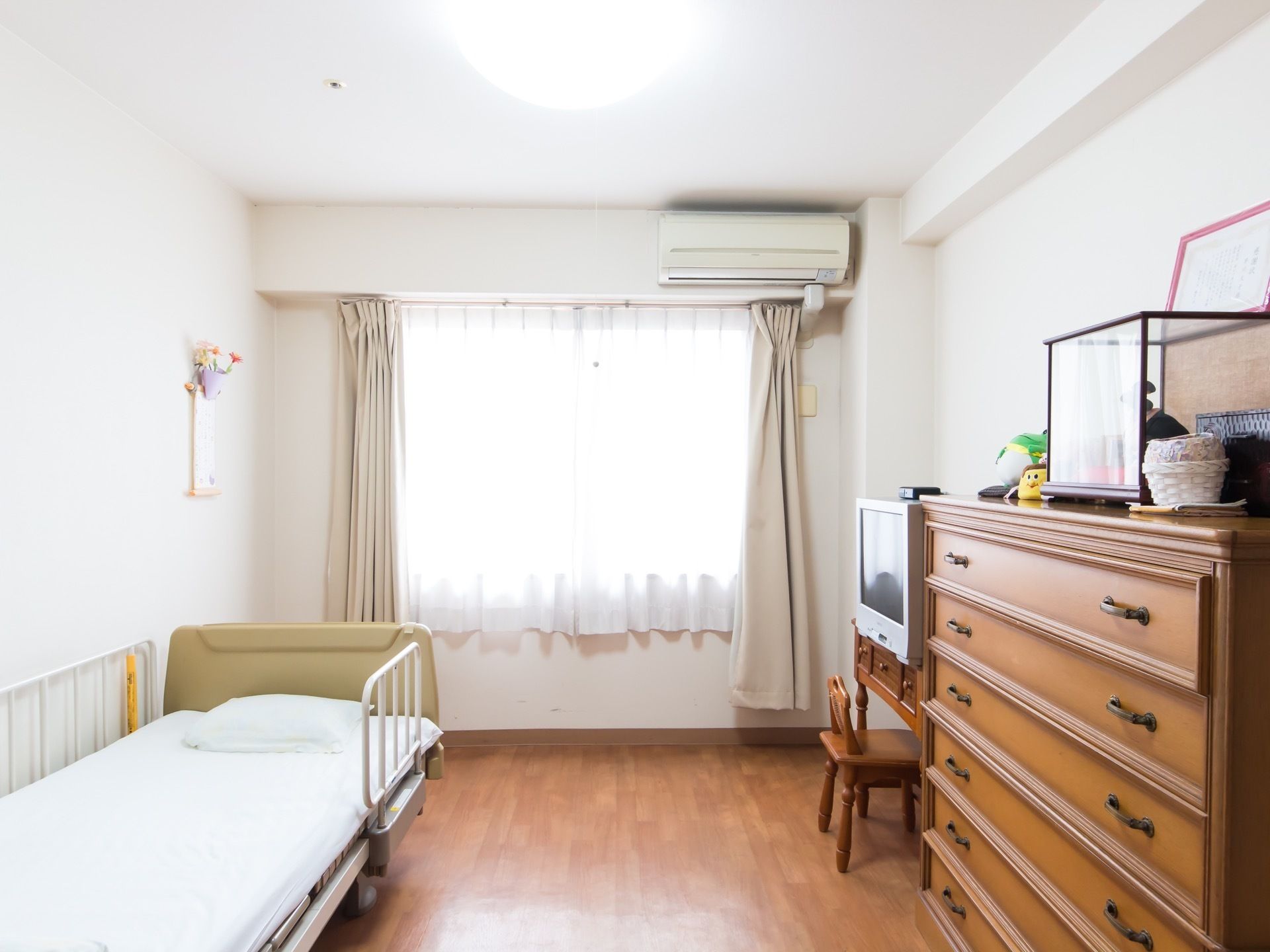 木目調の床と白い壁の居室がある。介護用ベッドがあり、箪笥、机、椅子が置かれている。箪笥の上にはガラスケースに入った人形や置物がある。