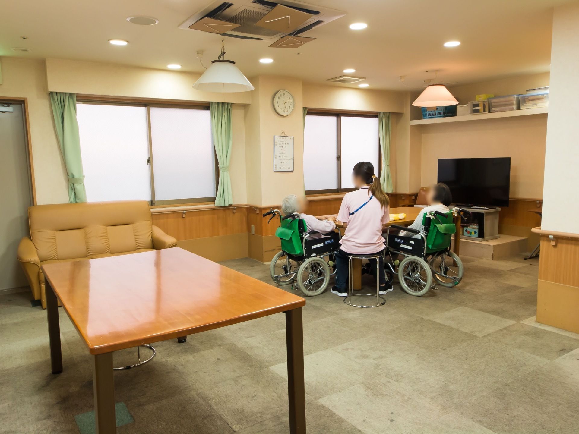 奥まった角部屋に、テーブルとソファが置かれている。窓が並んでおり、壁際には大きなテレビと棚がある。壁には手すりが設置されている。