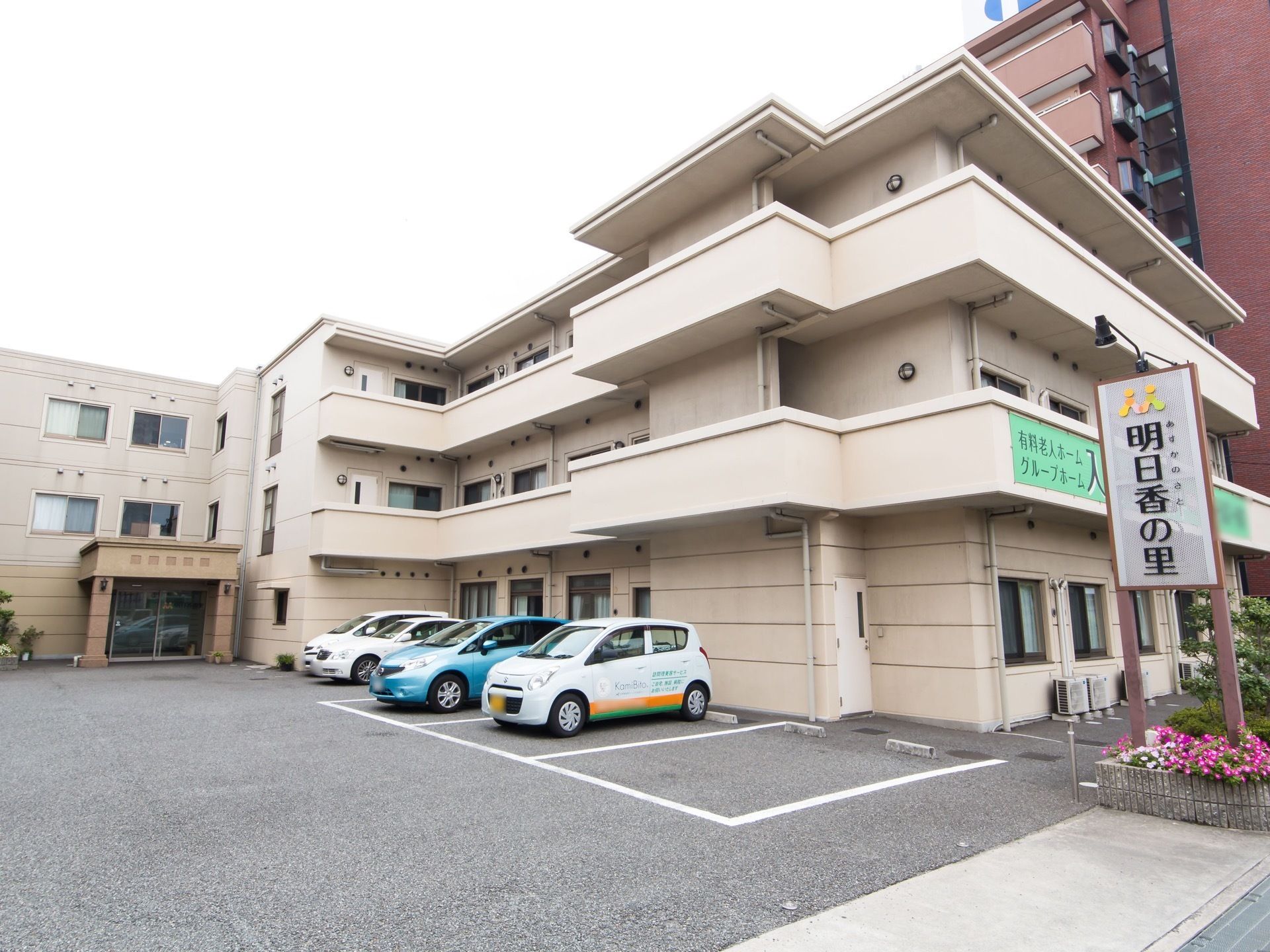 ベージュの壁の３階建ての大きな建物がある。建物に沿って駐車場がある。地面は舗装され、白線で駐車スペースが書かれている。