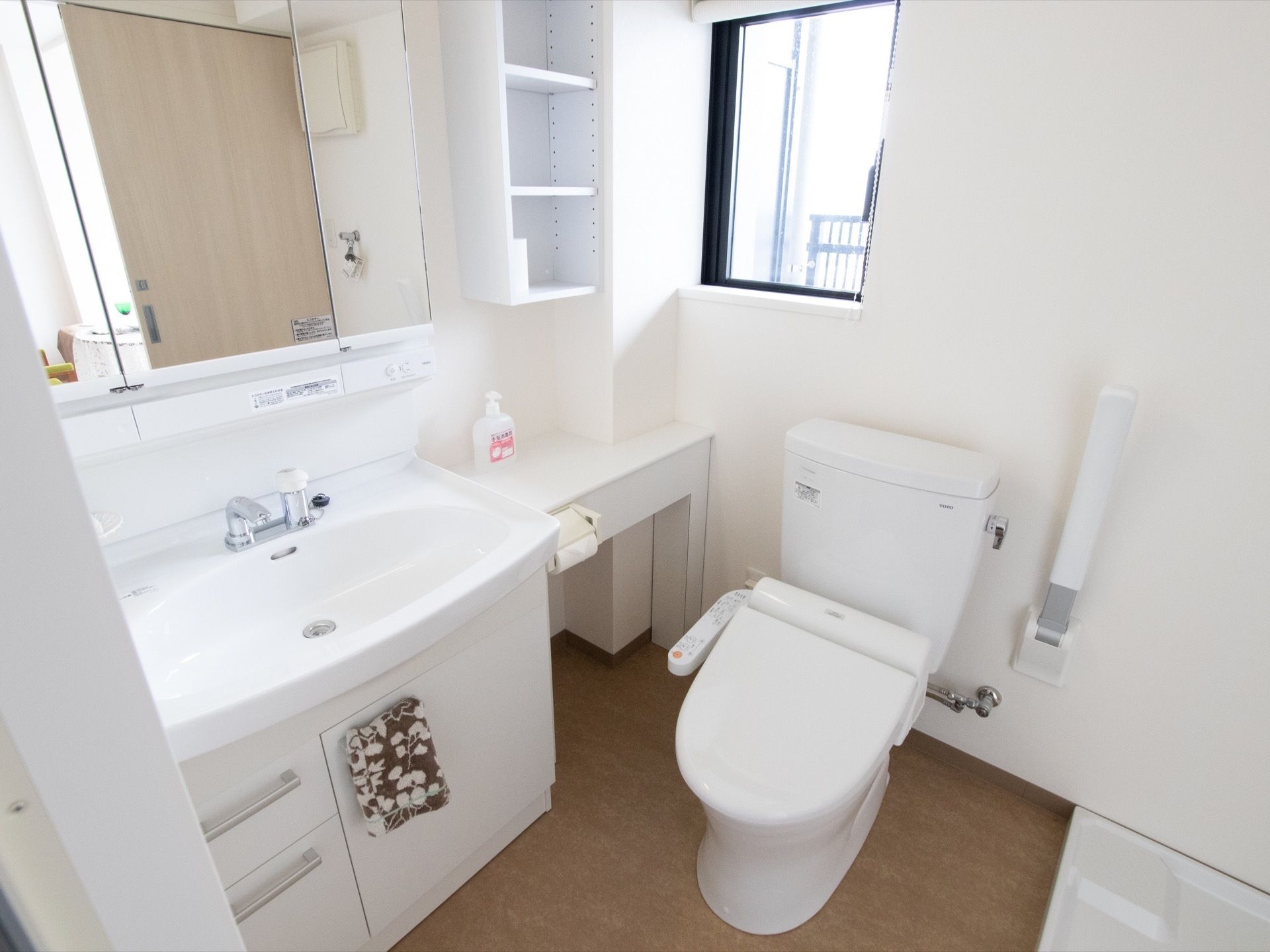 洗浄機付きの洋式トイレがある。横には可動式の手すりが取りつけられている。床面以外は白色で統一され、清潔感がある。