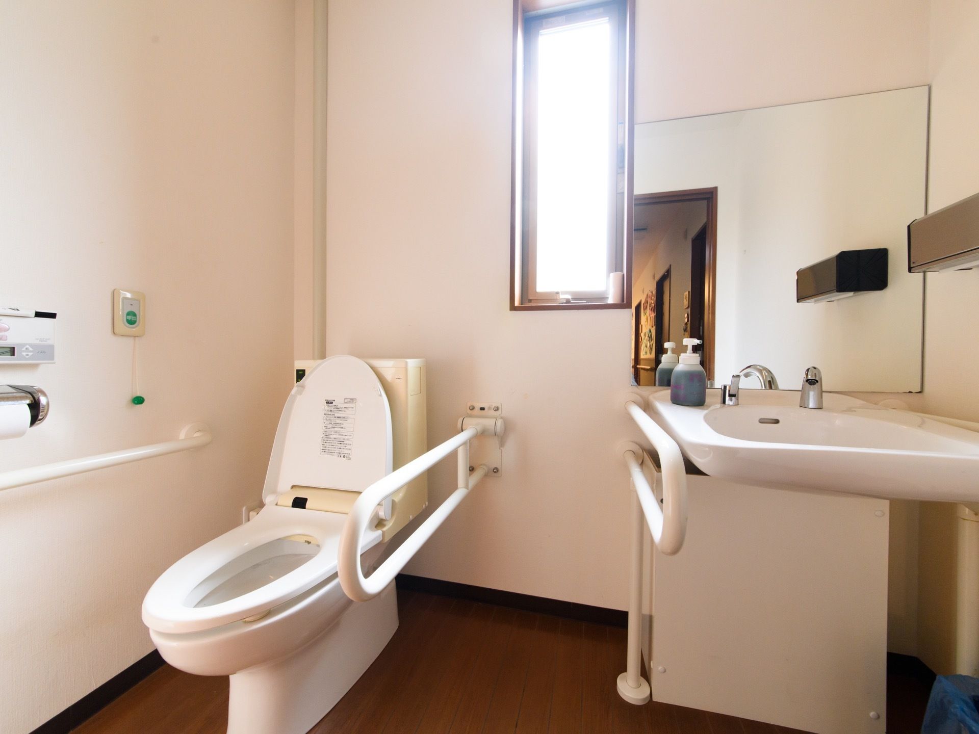 トイレには洋式便器設置されている。後方には縦長の窓があり、日が差し込んでいる。便器を手洗い器の横には手すりが取り付けられている。