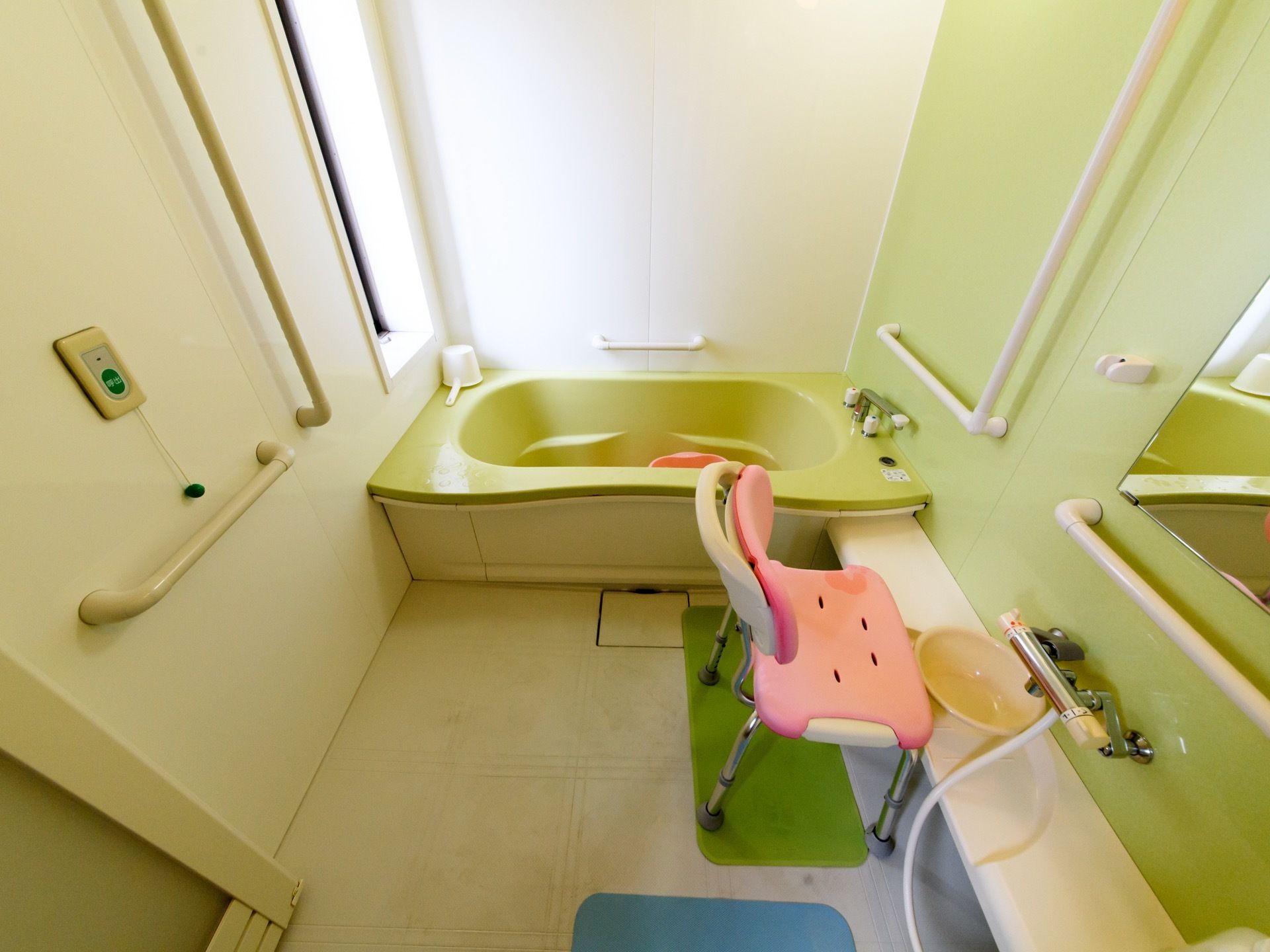浴室の洗い場には介護用お風呂椅子が置かれている。壁には呼び出しボタンがある。壁には縦型と横型の手すりが取り付けられている。
