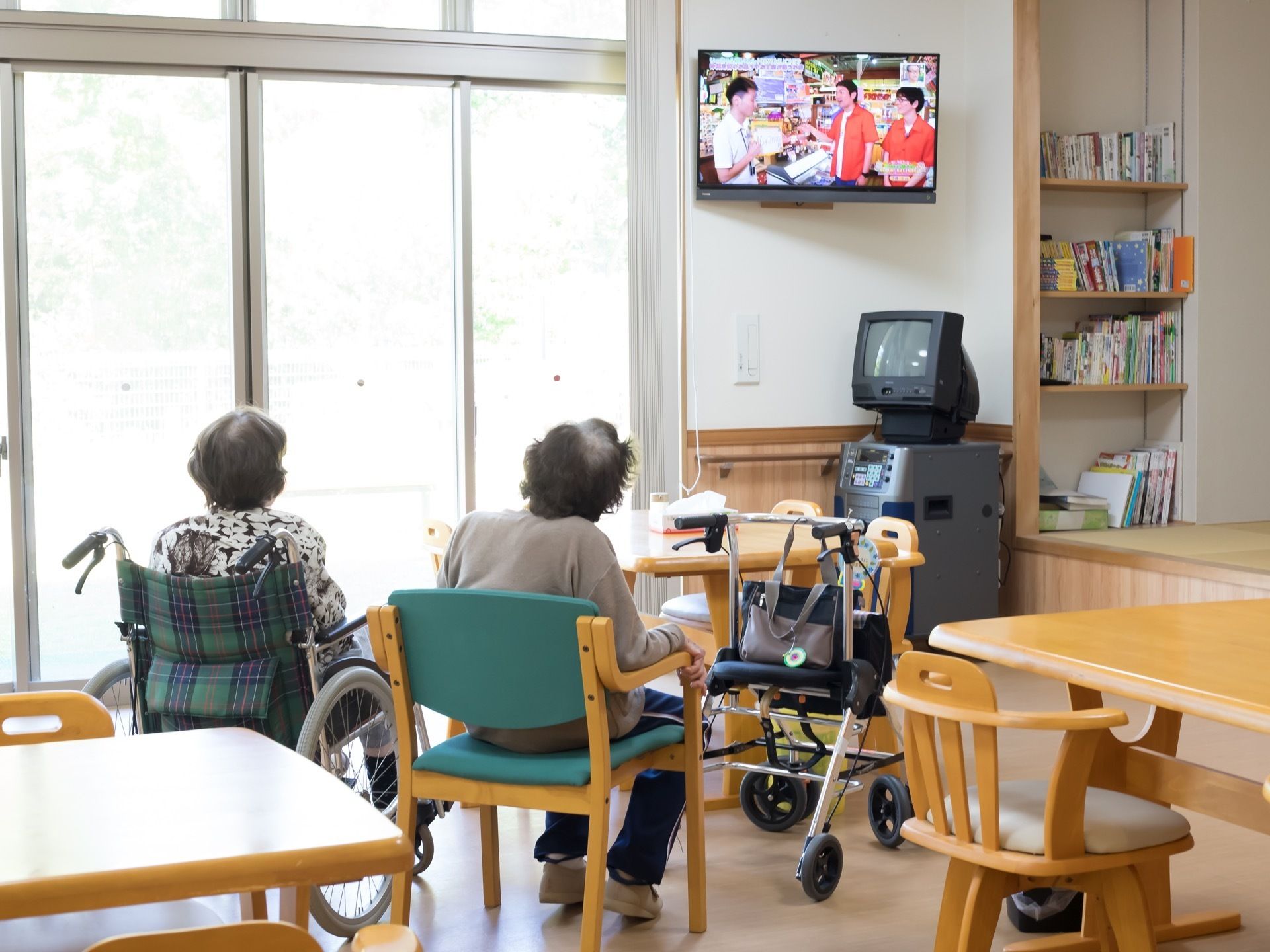 壁掛けテレビが設置されており、入居者様が椅子や車椅子に乗ったまま鑑賞している。本棚のスペースもあり、さまざまな書籍が置かれている。