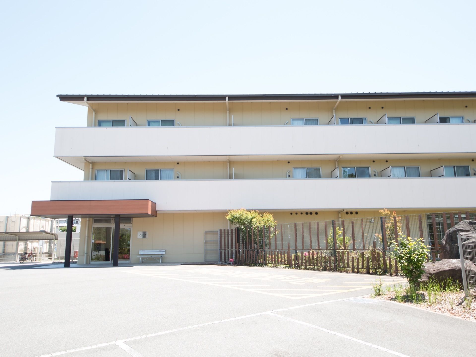 ３階建ての建物の入口前のスペースに平面駐車区画がある。入口には軒下があり、そばにはベンチも置かれている。