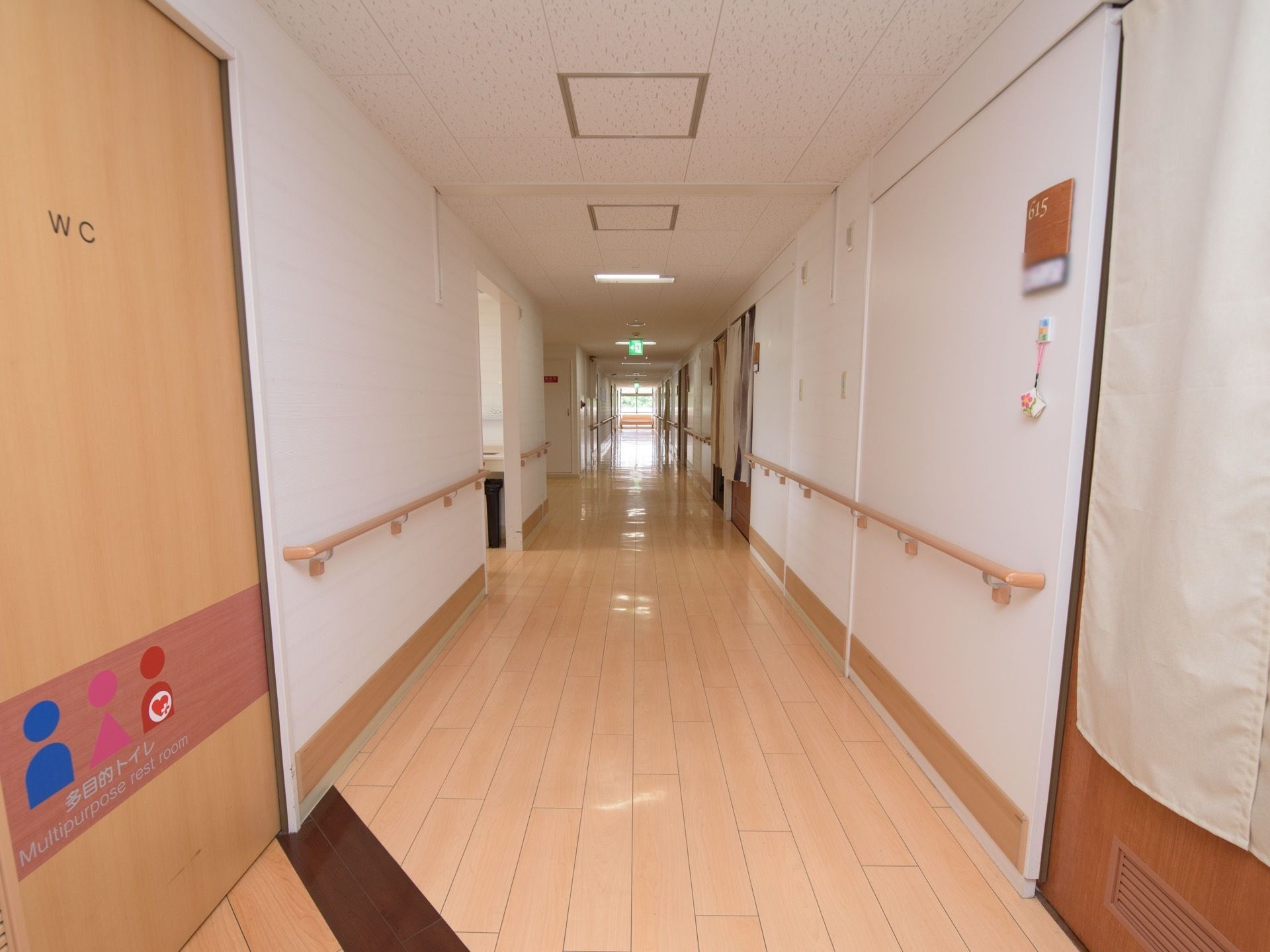 通路の床は光沢があるフローリングになっており、両側の壁には手すりが設置されている。左側には木目調のドアがあり、多目的トイレの表示がある。