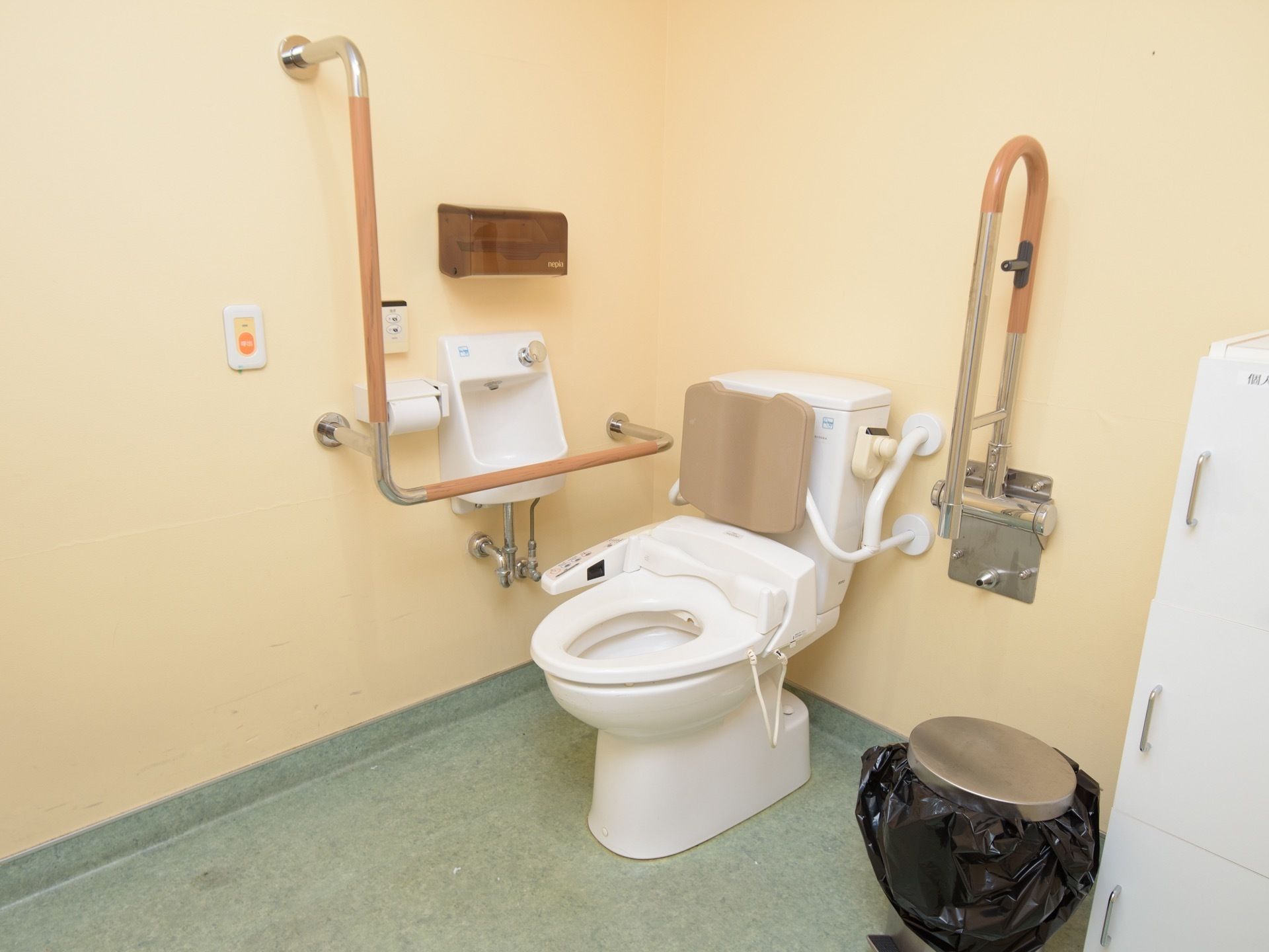 トイレは広々としており、温水洗浄便座が設置されている。壁にはＬ型や可動式の手すりが設置されていて、右側にはゴミ箱が置かれている。