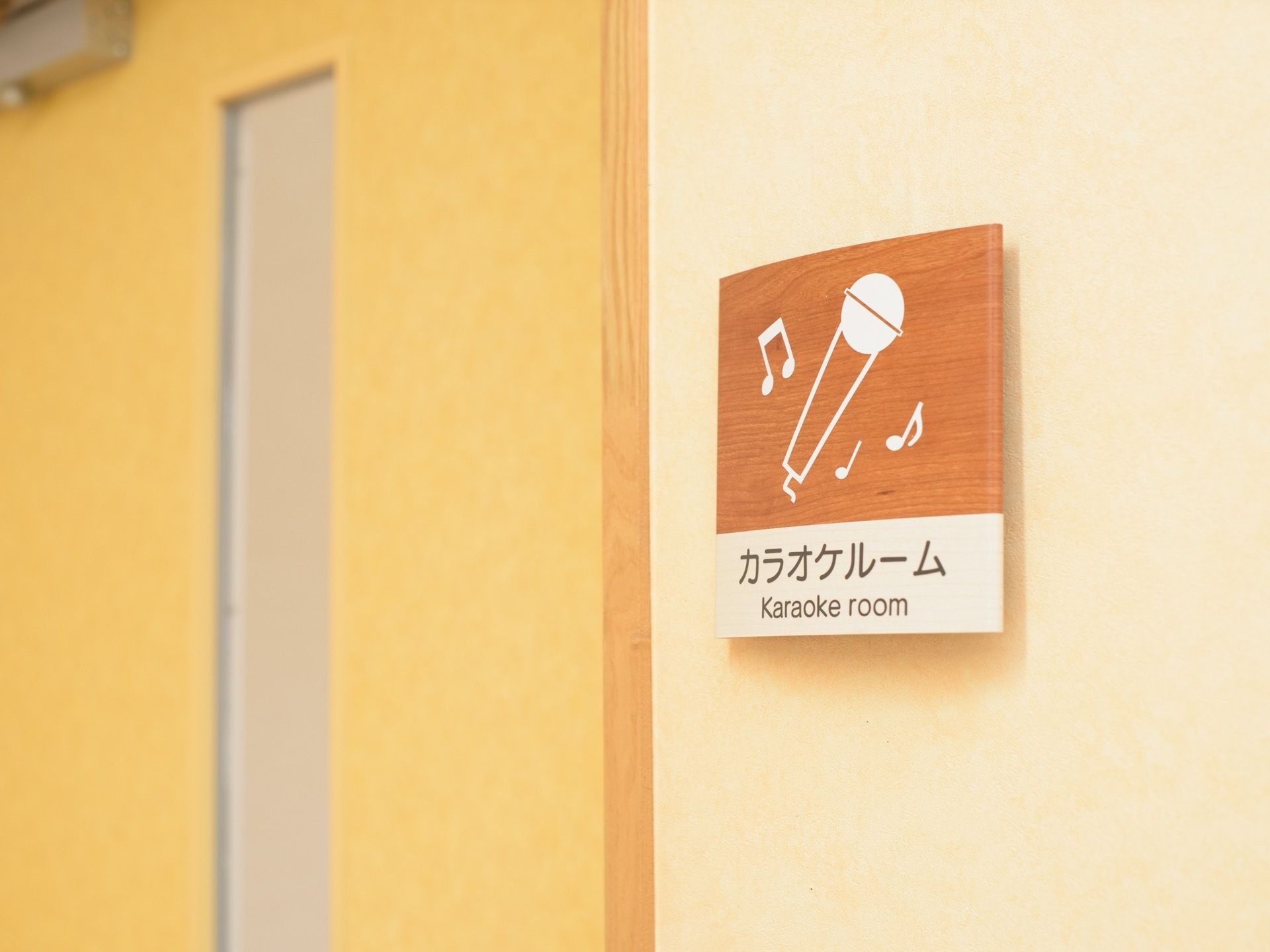 壁に、マイクの絵が描かれたカラオケルームのプレートが取り付けられている。側には細長い窓が付いたドアがある。