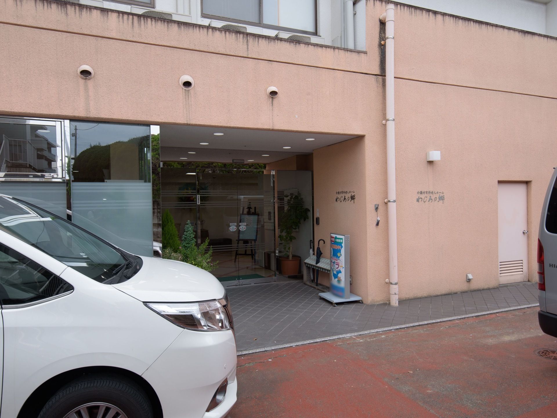 玄関の両脇には傘立てや植物が置かれており、入口は自動ドアになっている。壁には施設名が書かれていて、玄関の前には車が停められている。