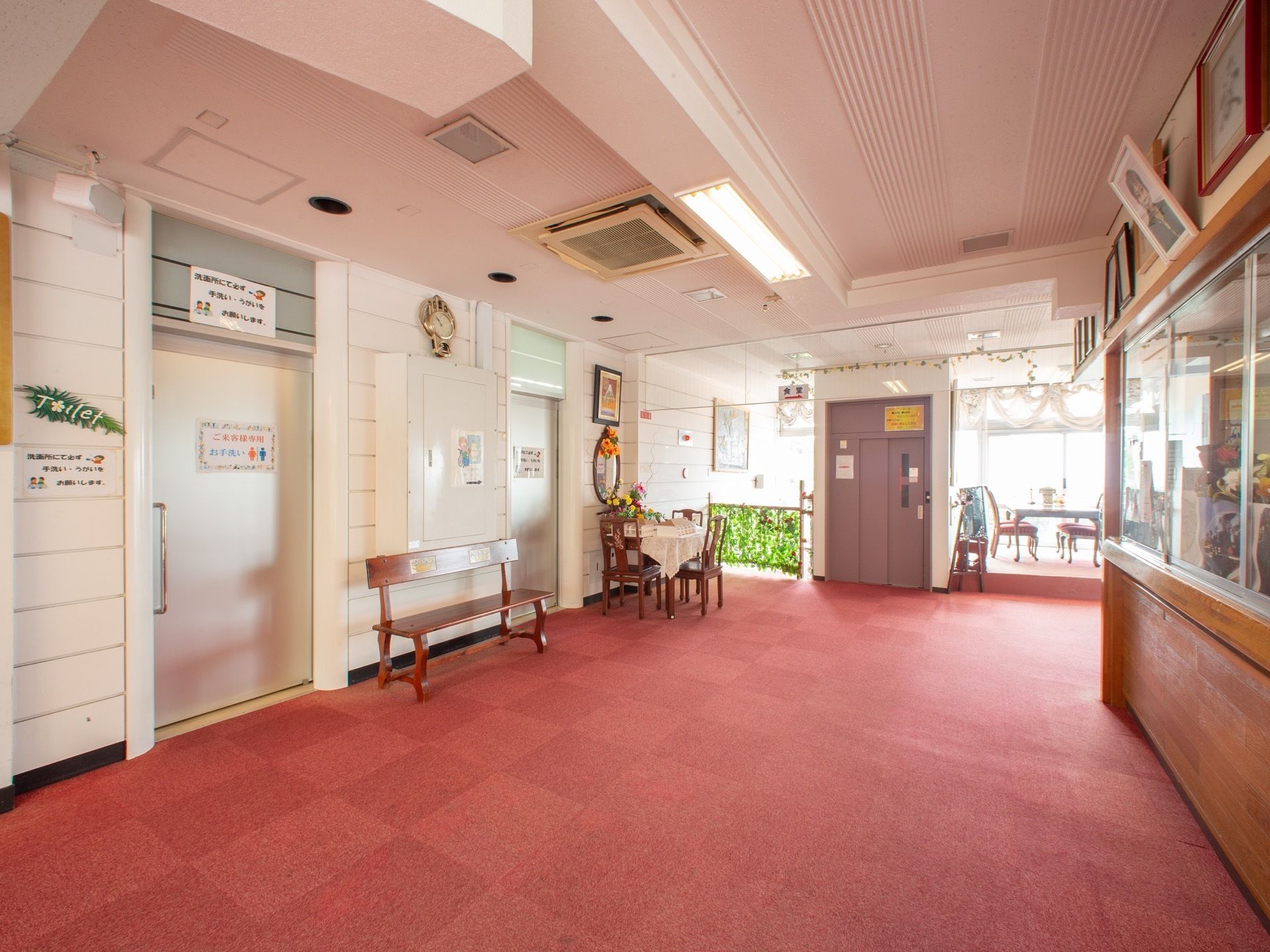 赤い床の空間に銀色のスライドドアのトイレが２か所ある。ベンチとテーブルと椅子が数脚置いてあり、灰色の扉のエレベーターがある。
