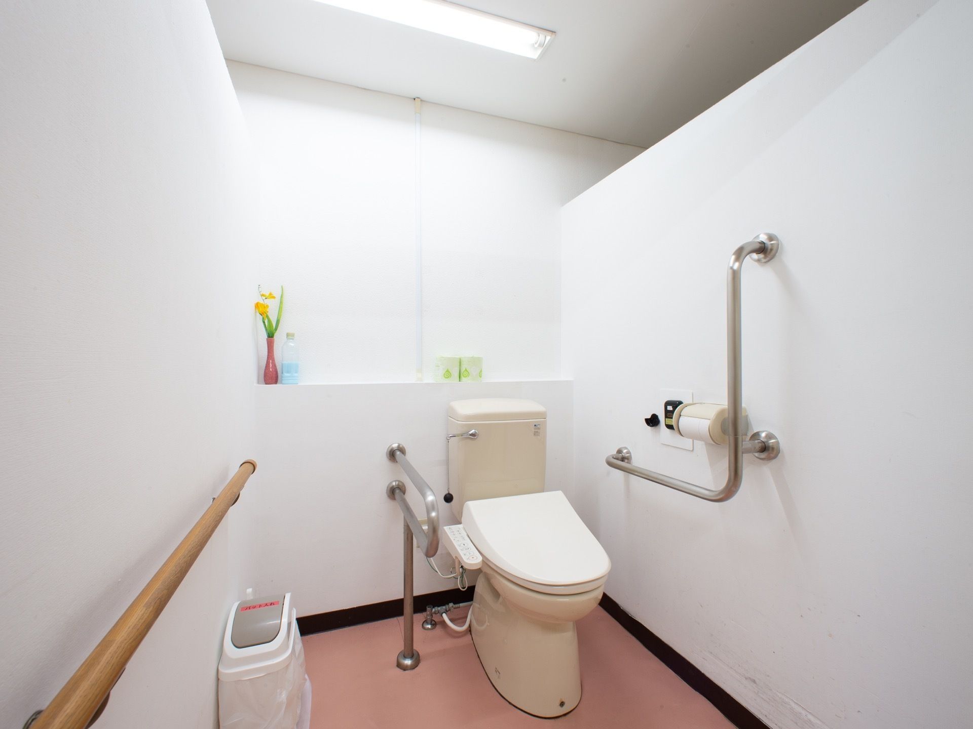 タンク付きのクリーム色の洋式トイレがあり、床に蓋付きのゴミ箱が置いてある。ピンク色の花瓶に黄色い花が飾られている。