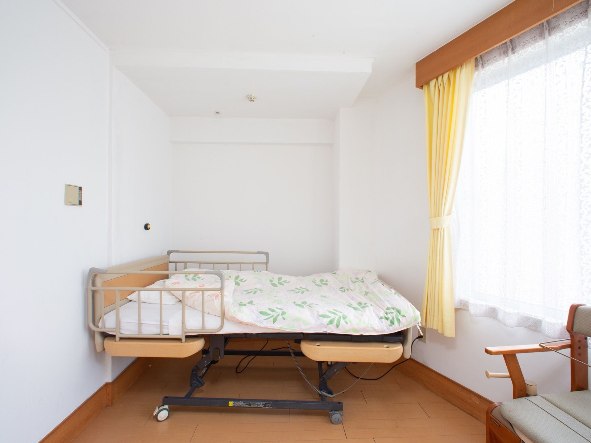 部屋の角に電動ベッドが置いてあり、柵が取り付けられている。クリーム色のカーテンが掛けられた窓の下に椅子が置いてある。