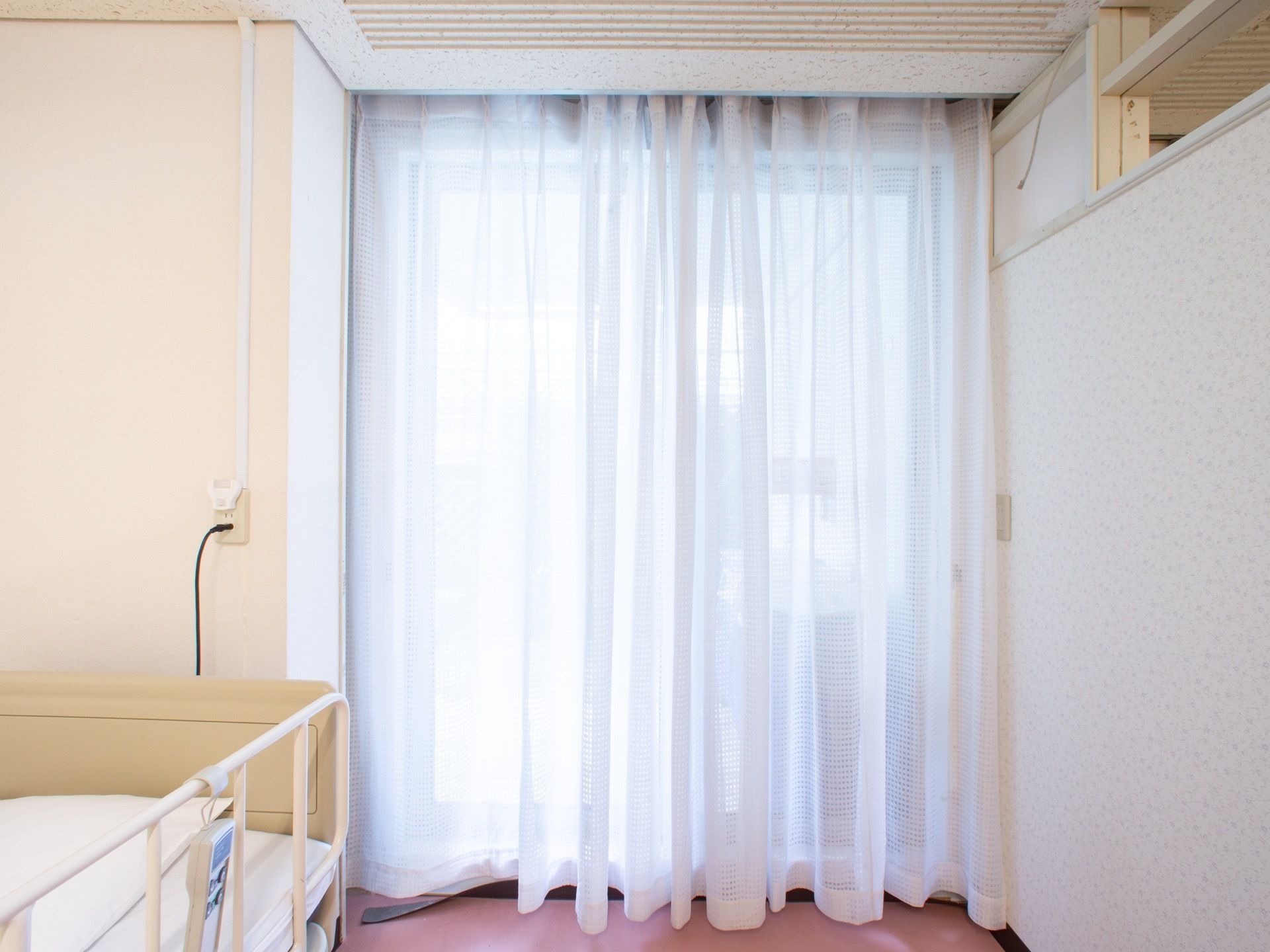 介護用ベッドのそばに高さのある窓が設置されている。白いレースのカーテンが引かれている。日中の陽射しを適度に調節できる。