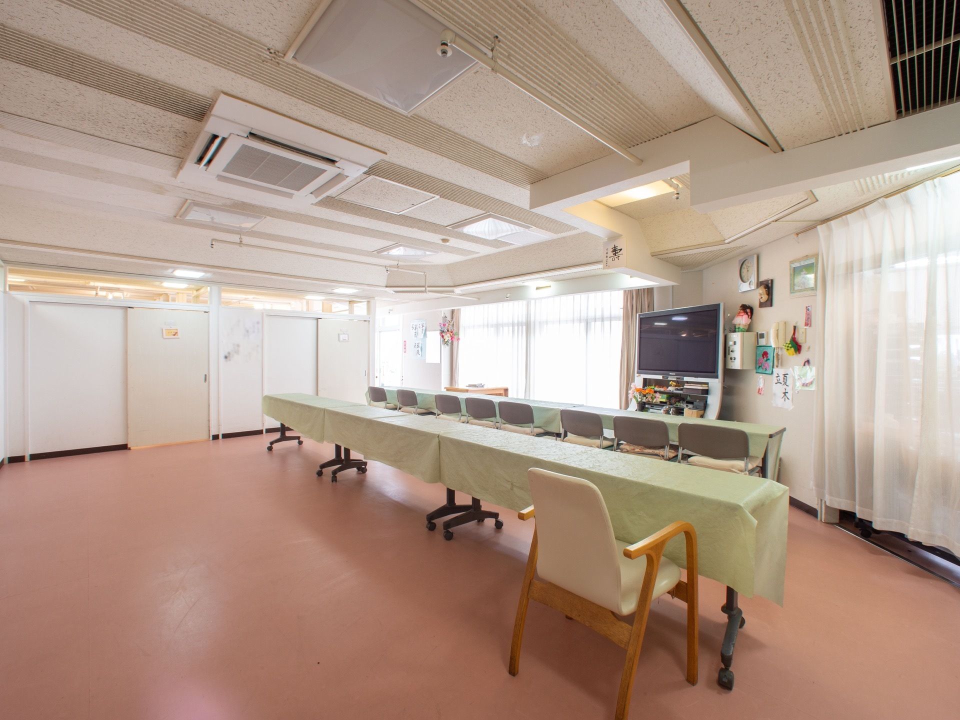 薄い緑色のテーブルクロスが掛けられた長机が並べて置いてある。壁際に設置されたテレビの両側に大きな窓がある。