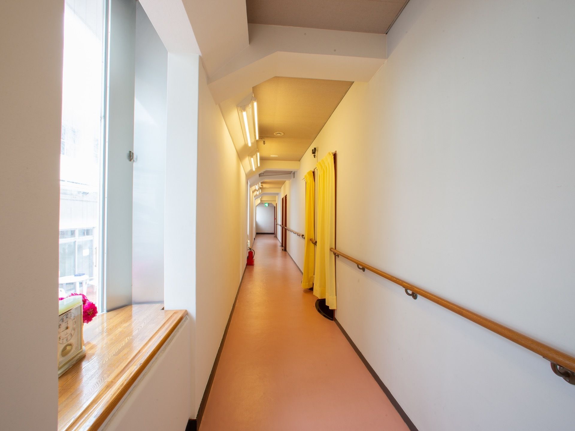 薄いオレンジ色の床の長い通路。腰高窓に時計が置いてある。クリーム色のカーテンが掛けられた入り口が２か所ある。