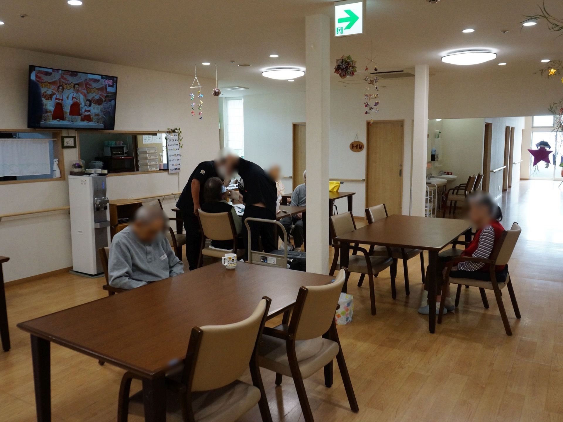 白い壁に茶色の木目調の床の広い共有スペース。壁に大型テレビが設置されていて、テーブルや椅子も複数備えられている。