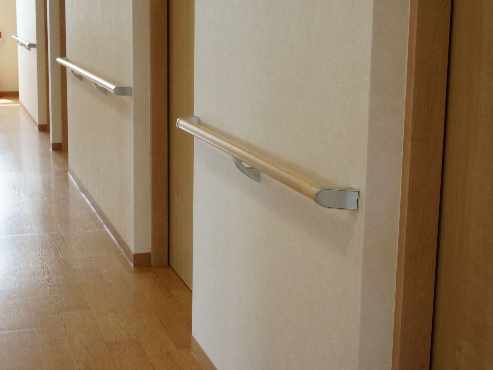 白い壁に茶色の木目調の床の通路。ドアがいくつか並んでいて、壁には茶色の木目柄の手すりが設置されている。