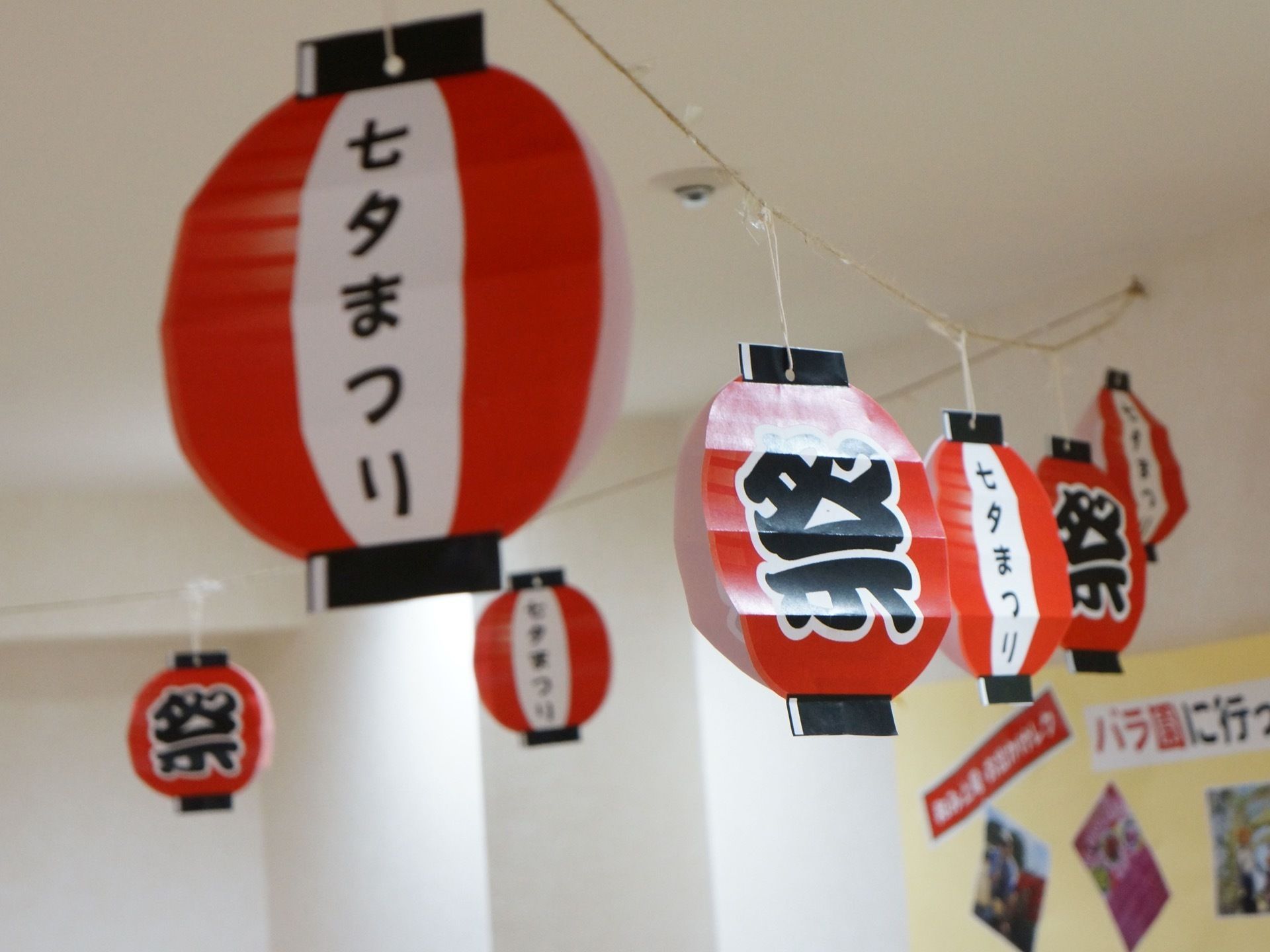 白色の天井からつるされた紐に、七夕まつりと書かれた赤と白と黒の提灯が飾りつけられている。また壁にはバラ園へ行った際の写真が飾られている。
