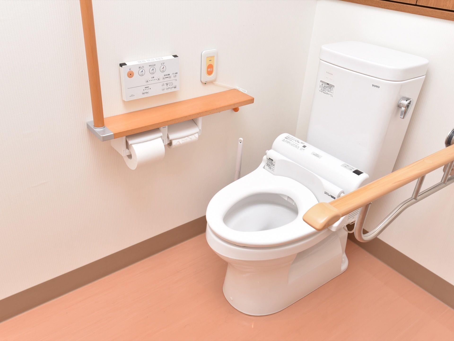 白色の壁と明るい茶系の床の内装に白色の便器を備えている。横の壁には、小さな棚があり、下には２つのトイレットペーパーホルダーがある。