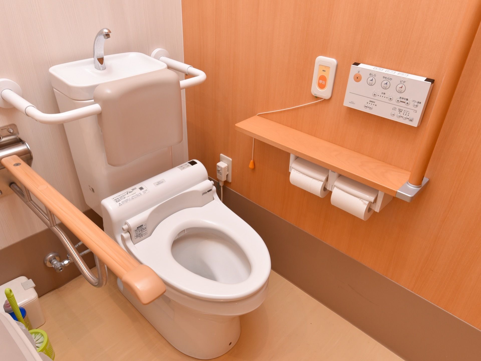 白色の壁に白色の便器を設置している。横の壁は木目調で、同色系の手すりと細長い棚がある。後部には、背もたれを備えている。