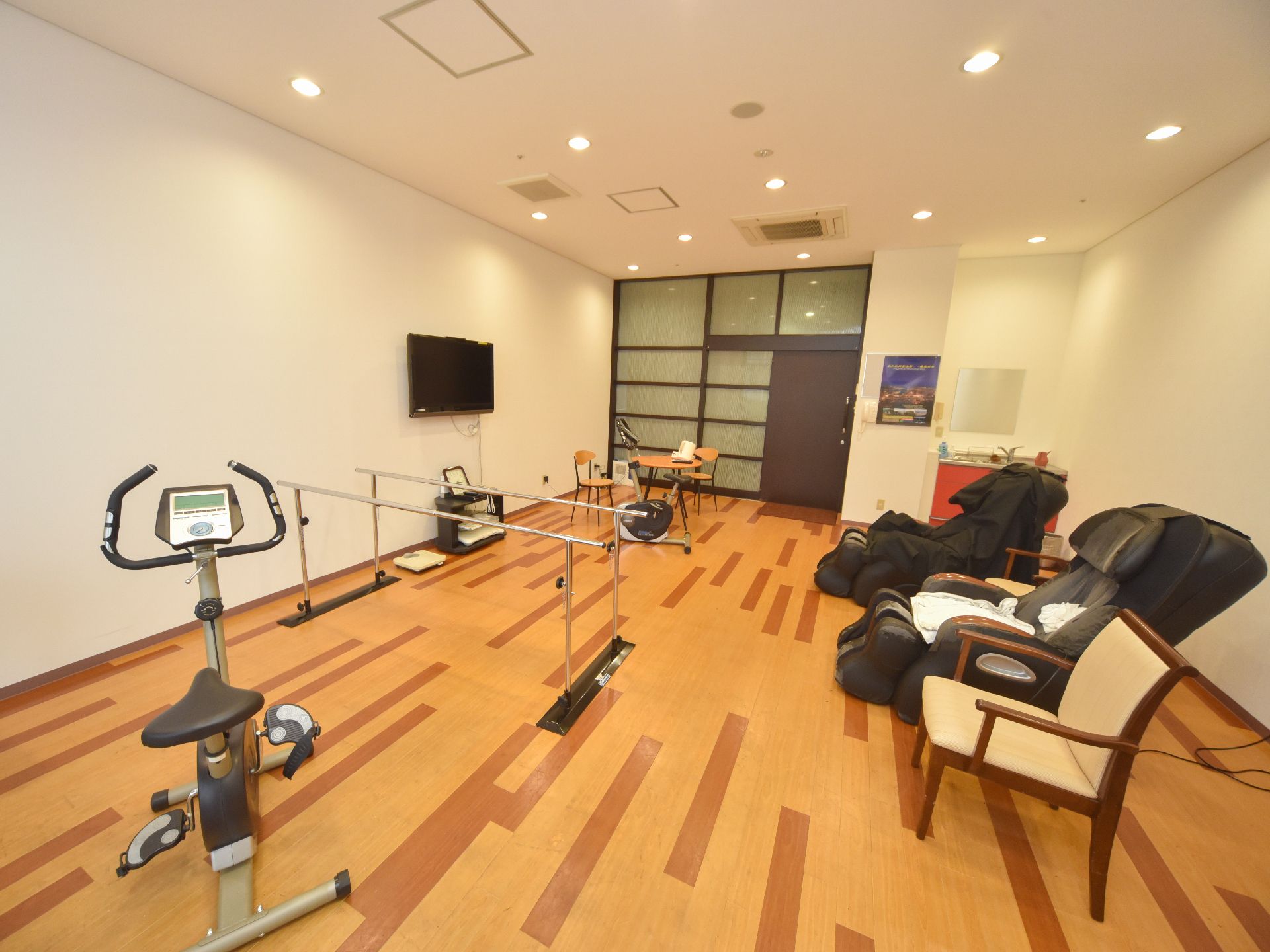 フラットなフロアに複数のトレーニング機器が置かれている。壁にはテレビが設置されており、向かい側の壁際にはマッサージチェアーがある。