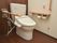 トイレの床はダークブラウンのフローリングで、背面の壁も同系色である。座って左側の白い壁にはナースコールが設置されている。