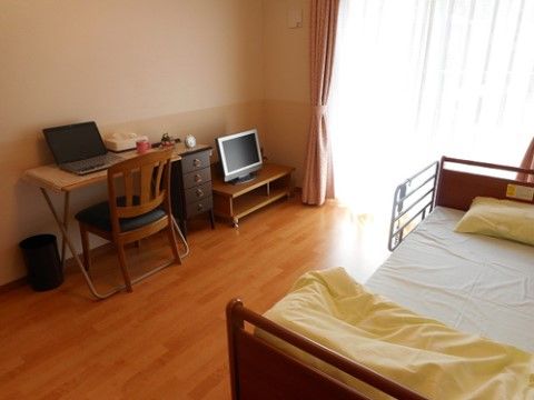 大きな窓から光が入る、あたたかな雰囲気の居室。転落防止柵のついたベッドとテレビ、椅子や机が置かれ、机の上にはパソコンがある。